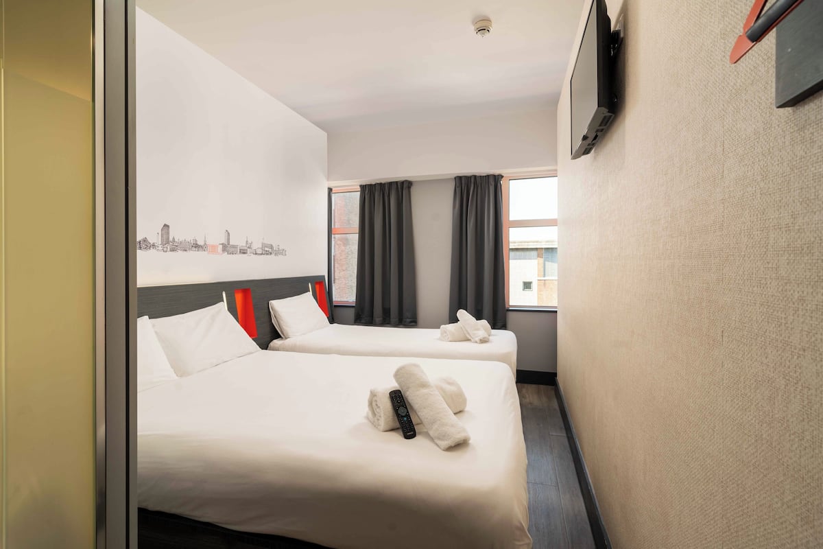 A hotel room with two beds, a cityscape artwork on the wall, a flat-screen TV, and a window with curtains.