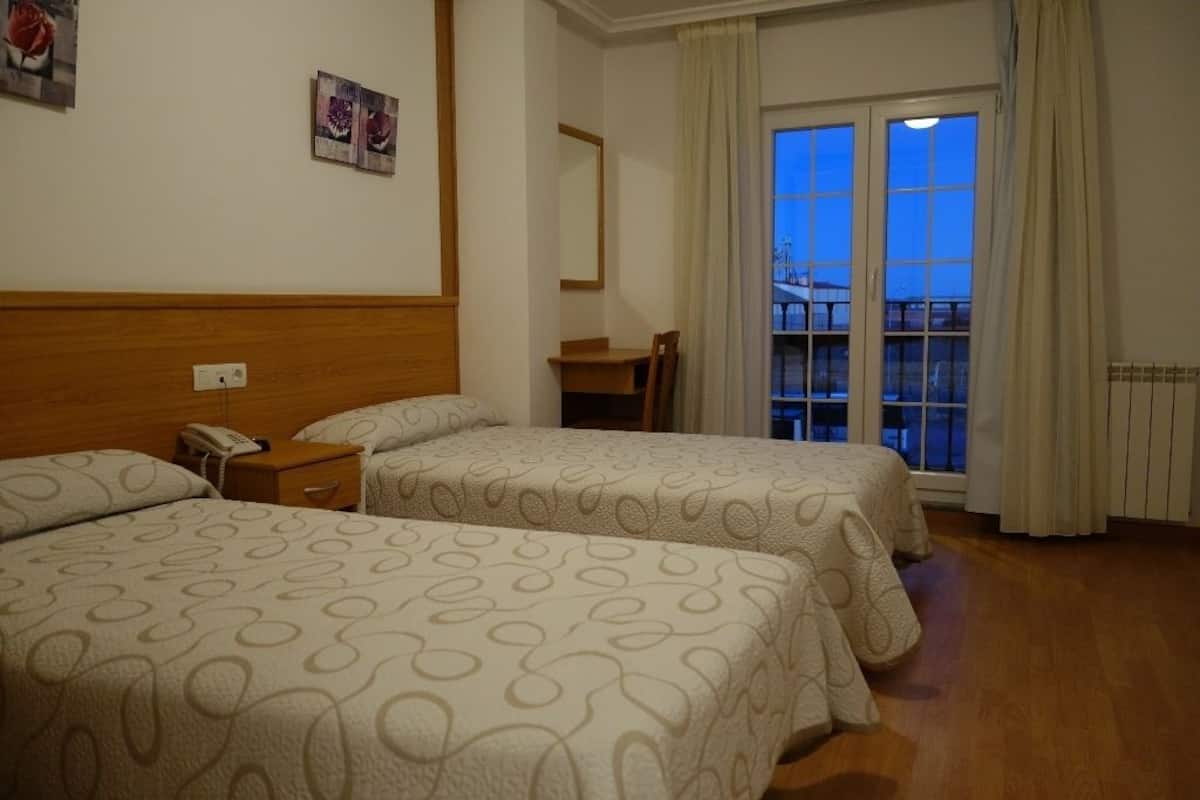 Quarto de hotel com duas camas, cabeceira de madeira, uma escrivaninha, uma cadeira e janela com cortinas.