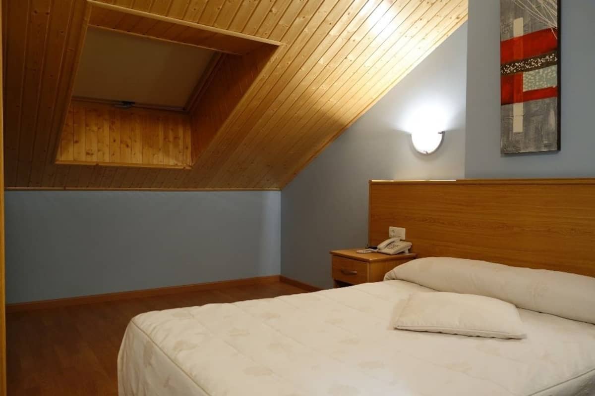 Quarto com teto inclinado, cabeceira de madeira, mesa de cabeceira com telefone e cama com almofadas.