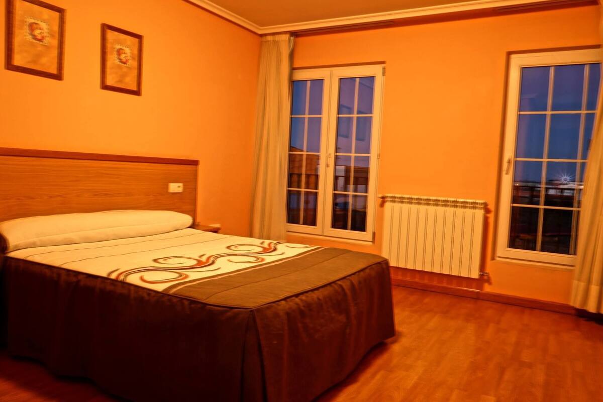 Quarto com cama, duas janelas, radiador e cabeceira de madeira.