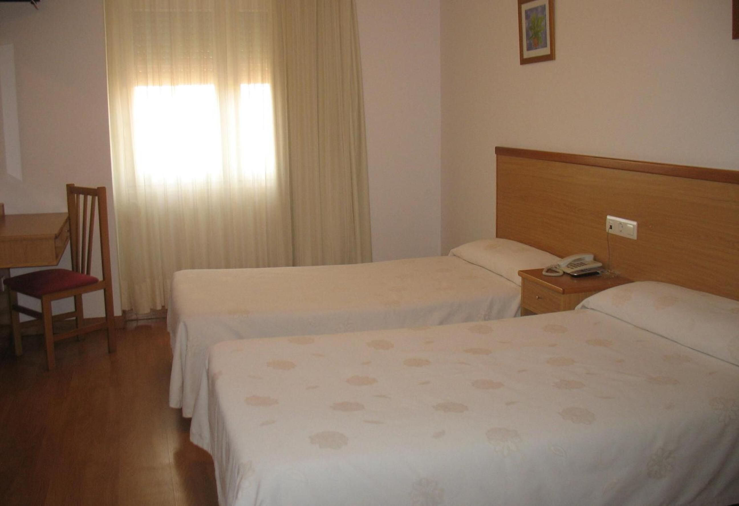 Hotelkamer met twee bedden, een houten hoofdbord, een nachtkastje met telefoon, een stoel met een rode kussen en een raam met dunne gordijnen.