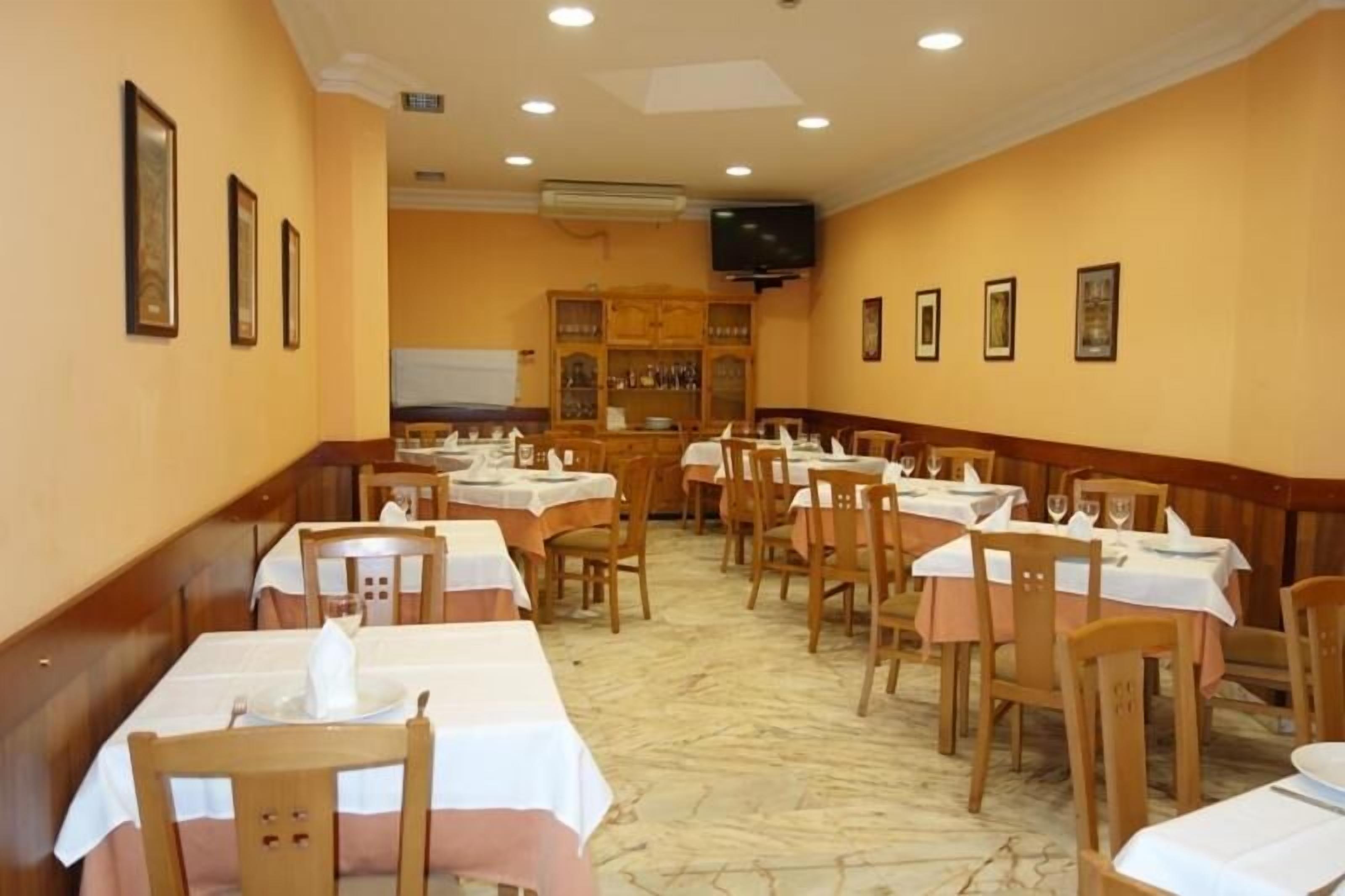 A restaurant with multiple tables and chairs, a television mounted on the wall, and a wooden cabinet with glass doors in the background.