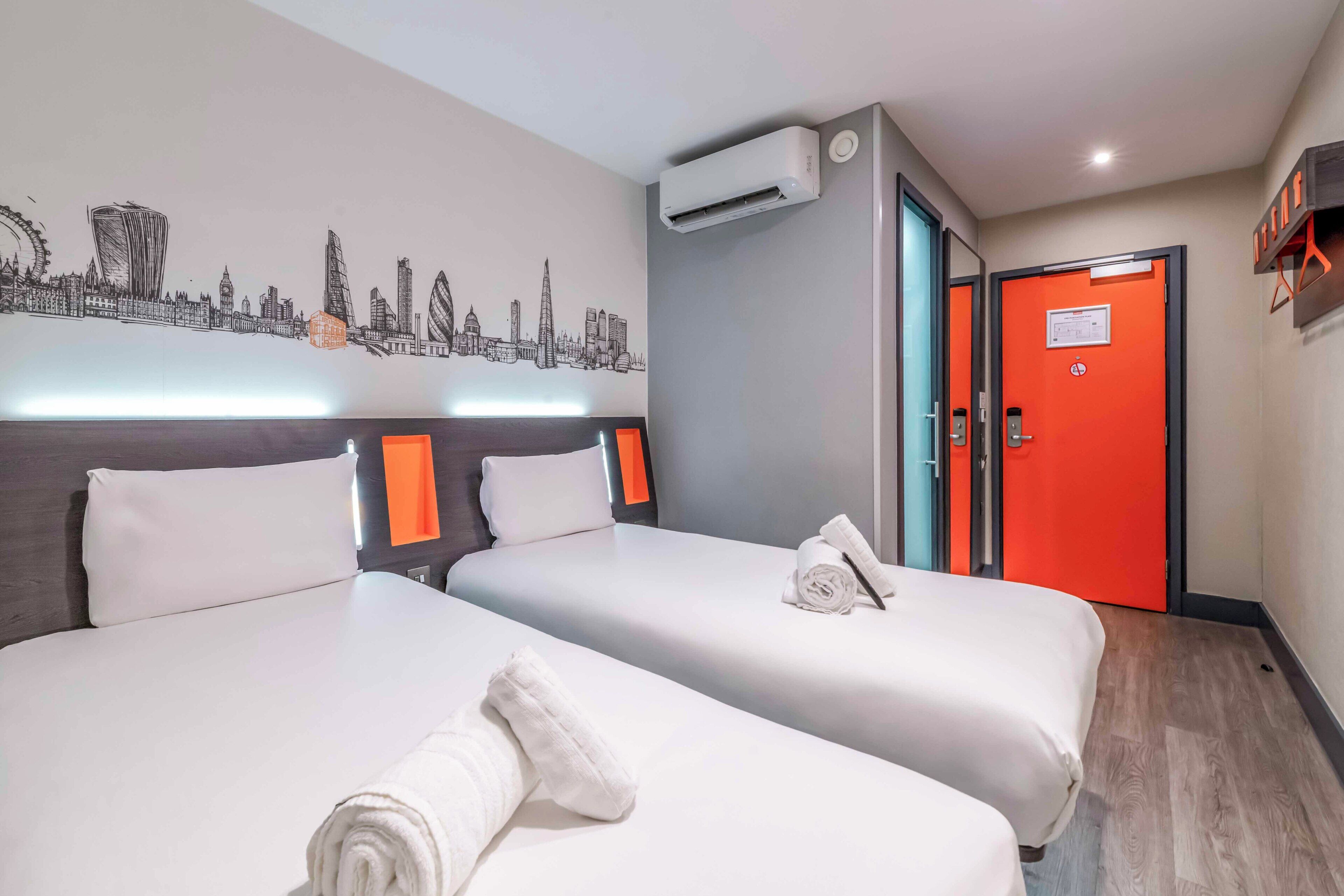 A hotel room with two beds, a cityscape wall art, an air conditioning unit, and a bright red door.