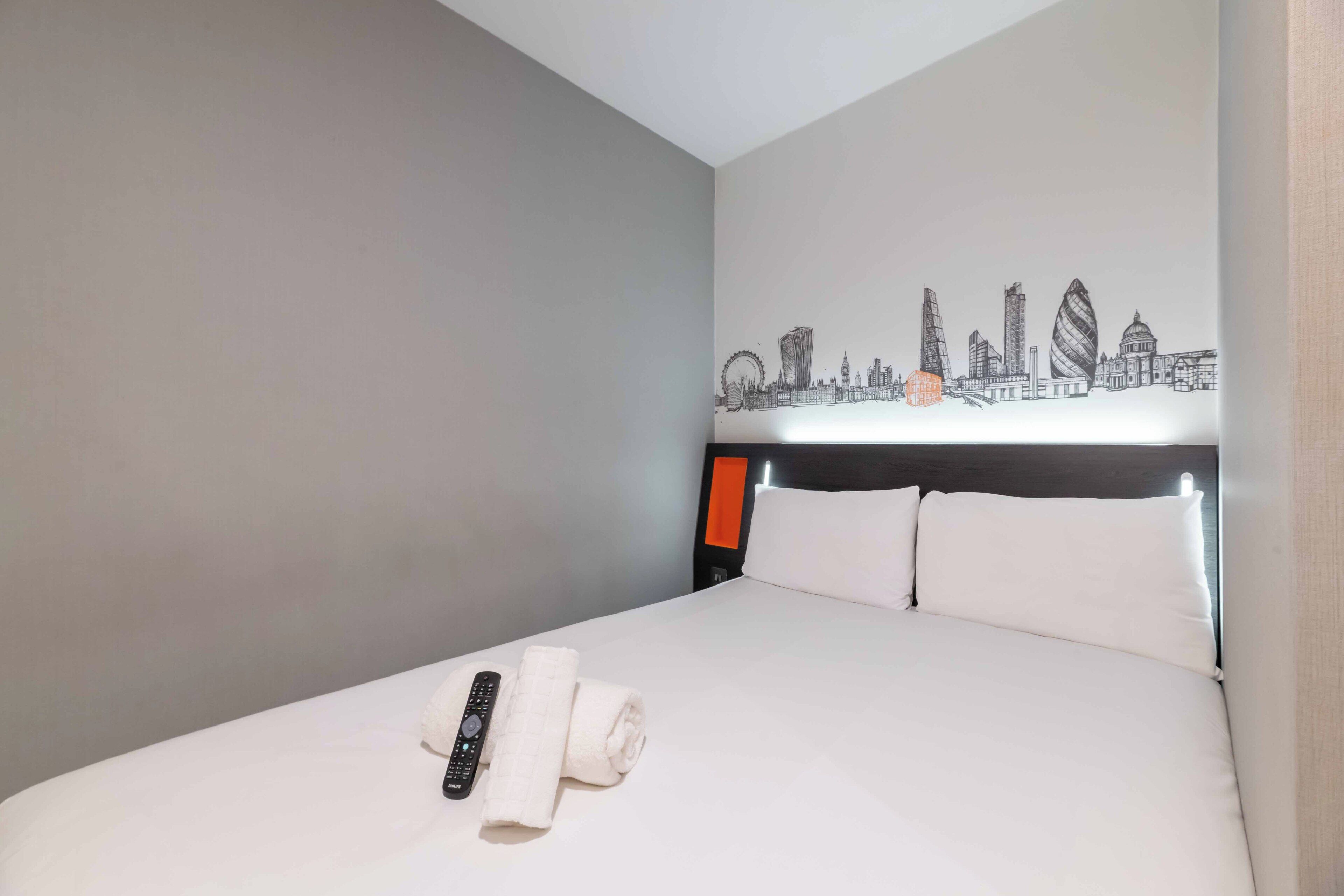 A hotel room with a bed, a cityscape wall art, a remote control, and a rolled towel.