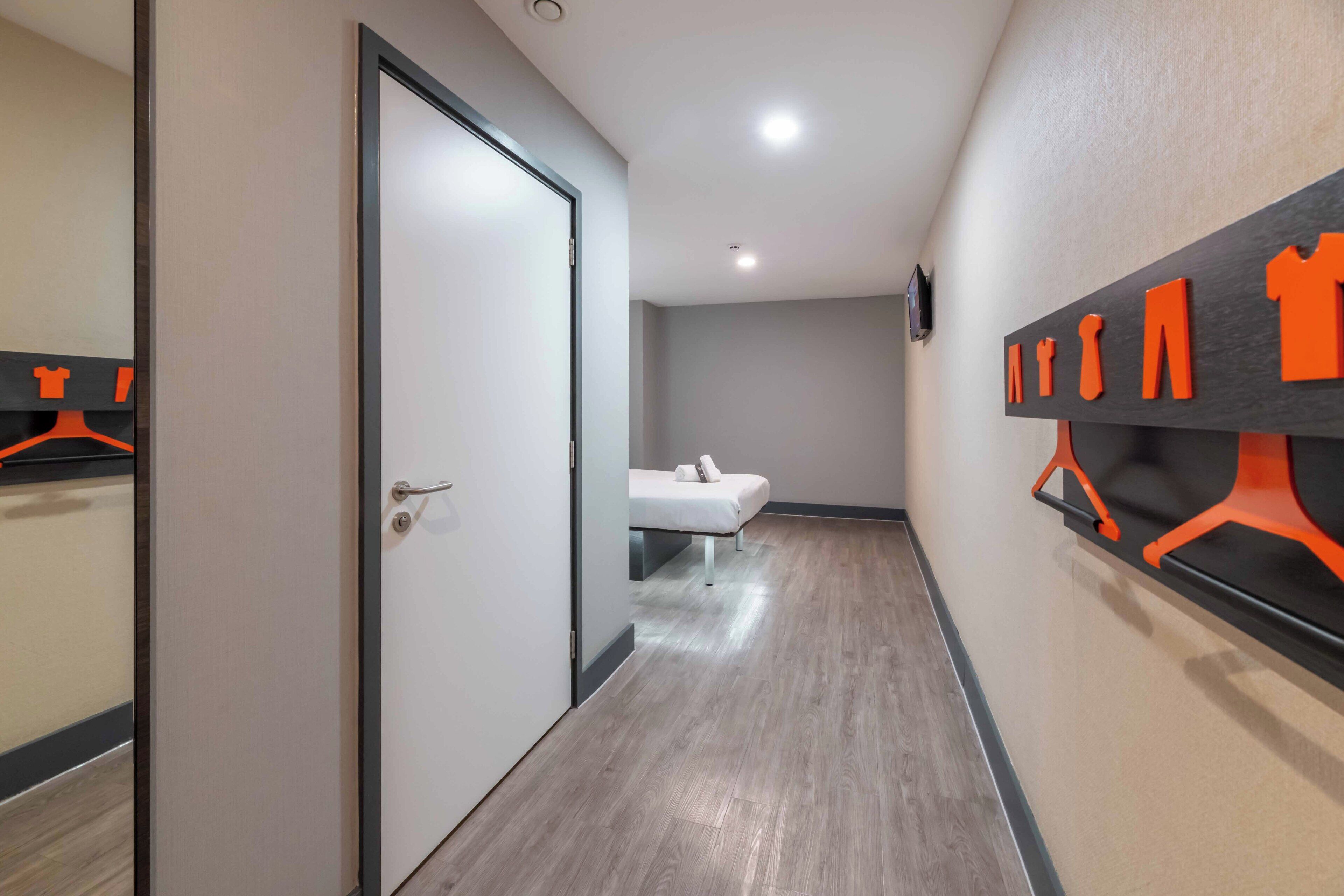 A hotel corridor with a bed, a door, and a wall with orange hangers.