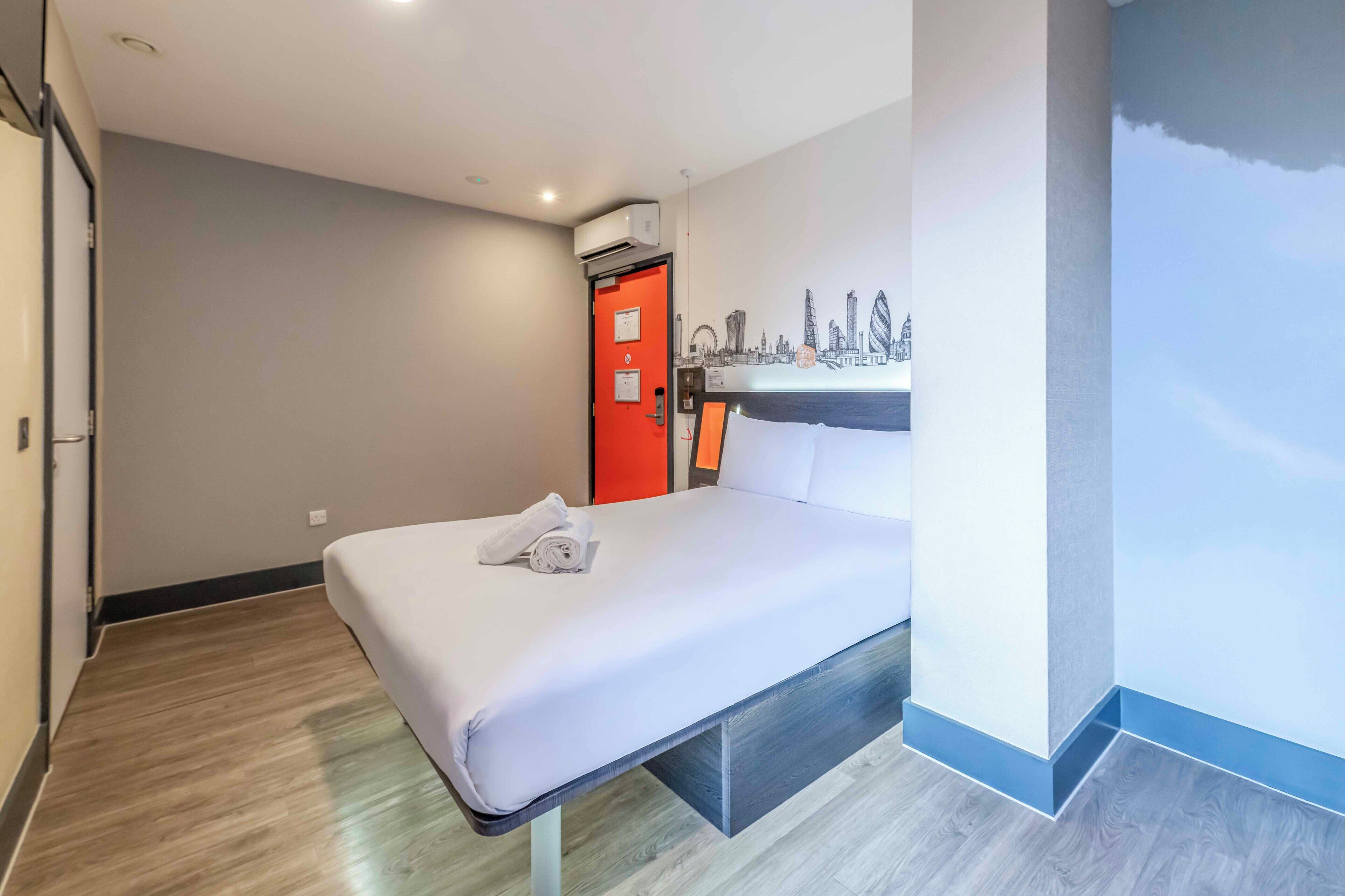 Une chambre d’hôtel avec un lit, une porte rouge, un climatiseur fixé au mur et une fresque représentant un paysage urbain.