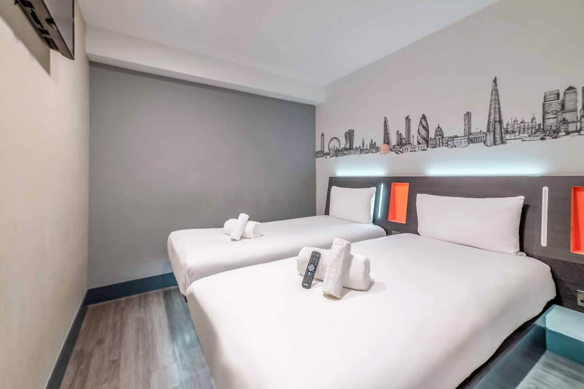 A hotel room with two beds, a cityscape wall art, and a remote control on the beds.