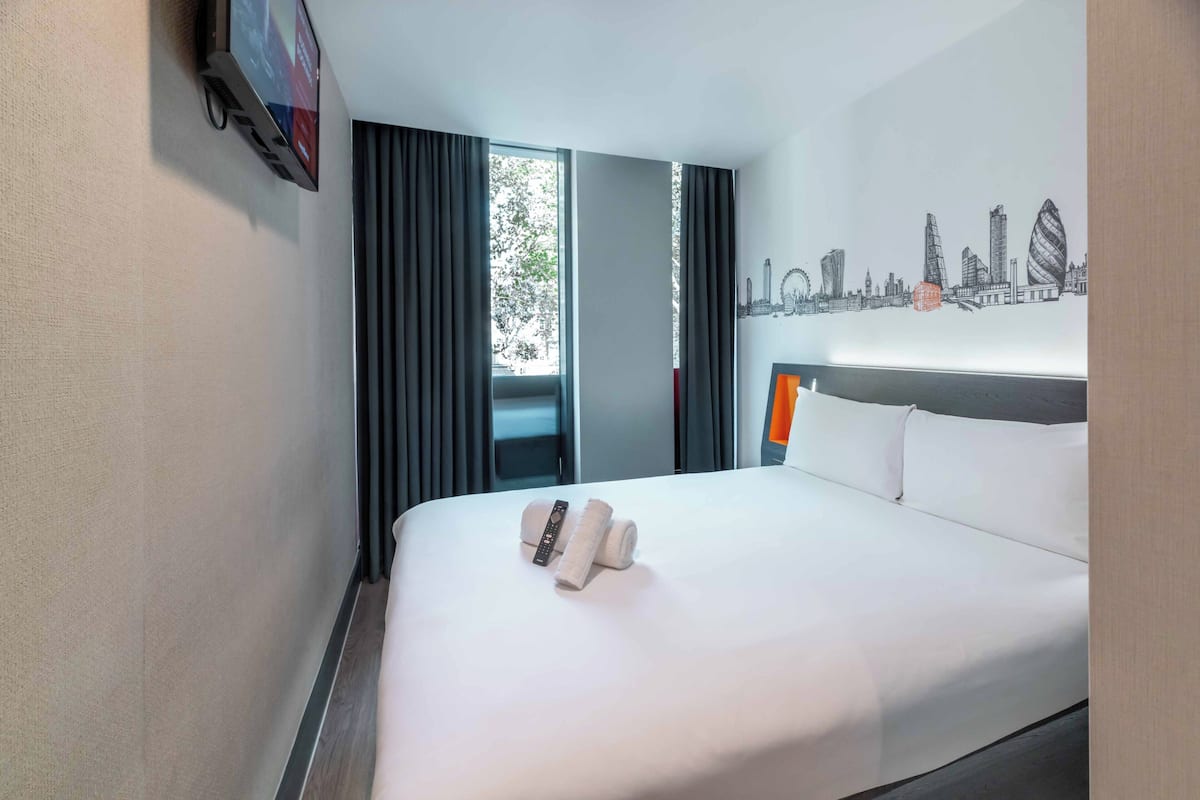 A hotel room with a large bed, a cityscape wall art, a TV, and a window with curtains.