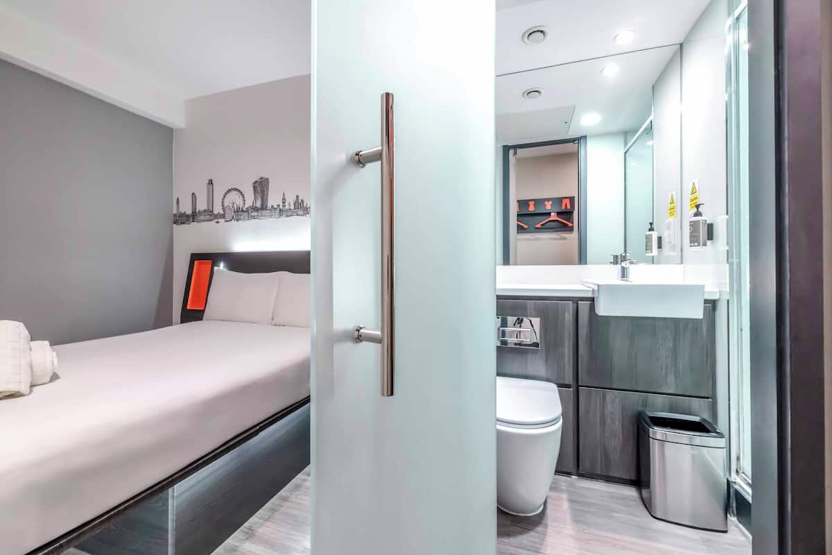 A modern hotel room with a bed, a cityscape wall art, a bathroom with a toilet, sink, and trash bin.