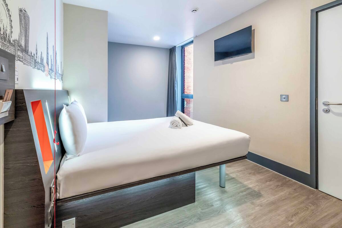 A hotel room with a bed, a flat-screen TV, a window, and a door.
