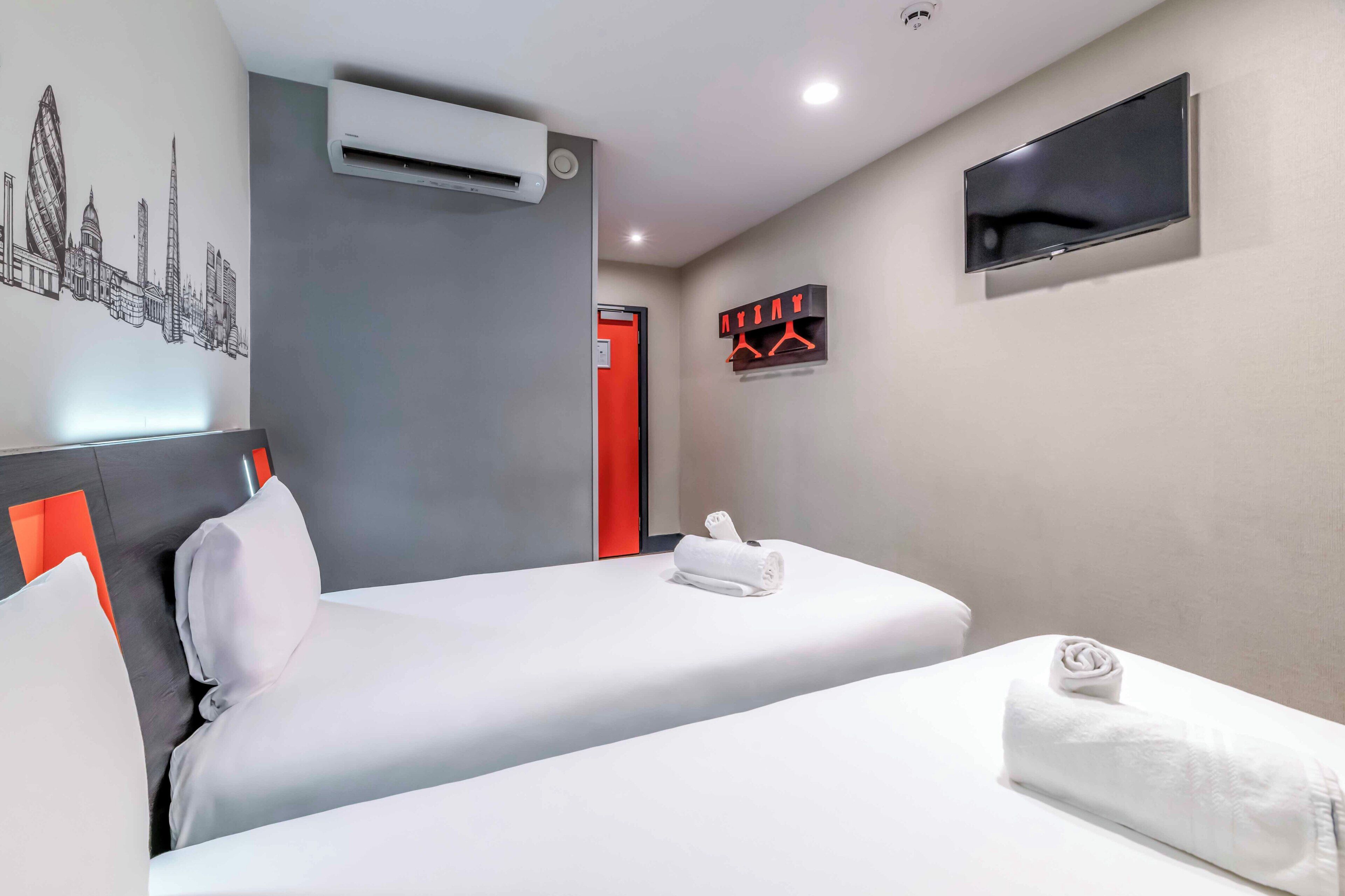 A hotel room with a bed, pillows, a flat-screen TV, a wall-mounted air conditioner, and a red door.