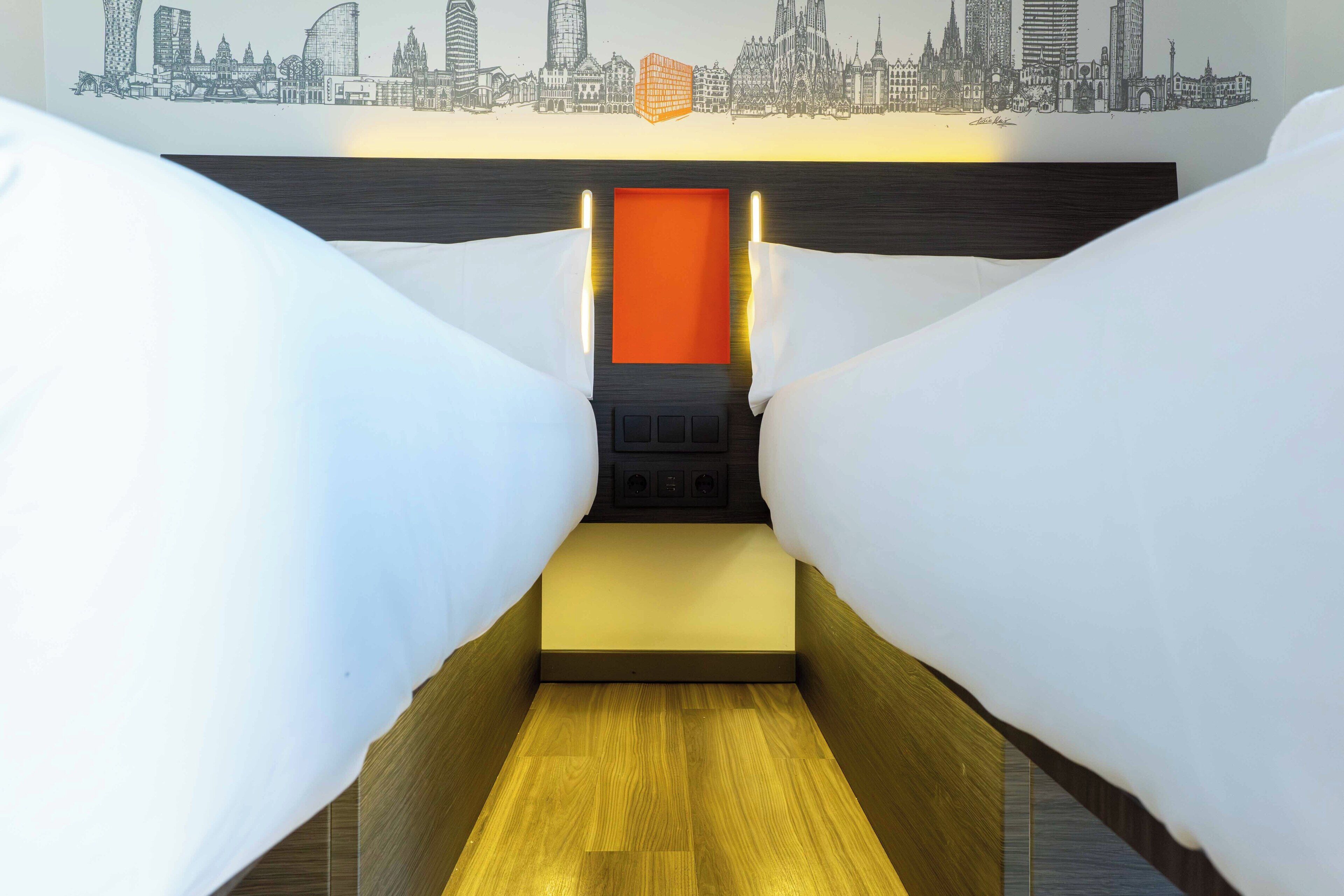 Une pièce étroite et exiguë, avec un plancher en bois, un lit doté d’un matelas jaune et une fresque représentant un paysage urbain sur le mur.