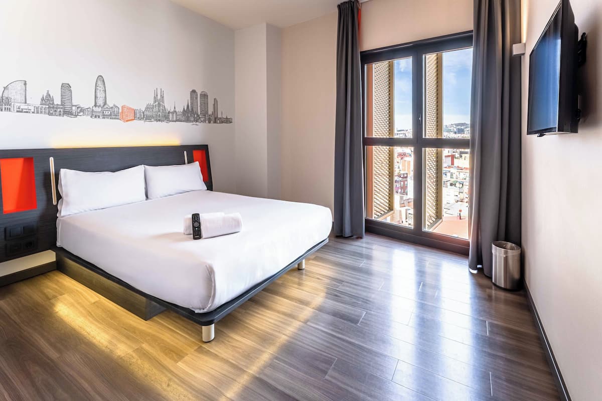 Habitación de hotel con una cama, un cuadro de arte urbano en la pared, un televisor y un ventanal con vistas.