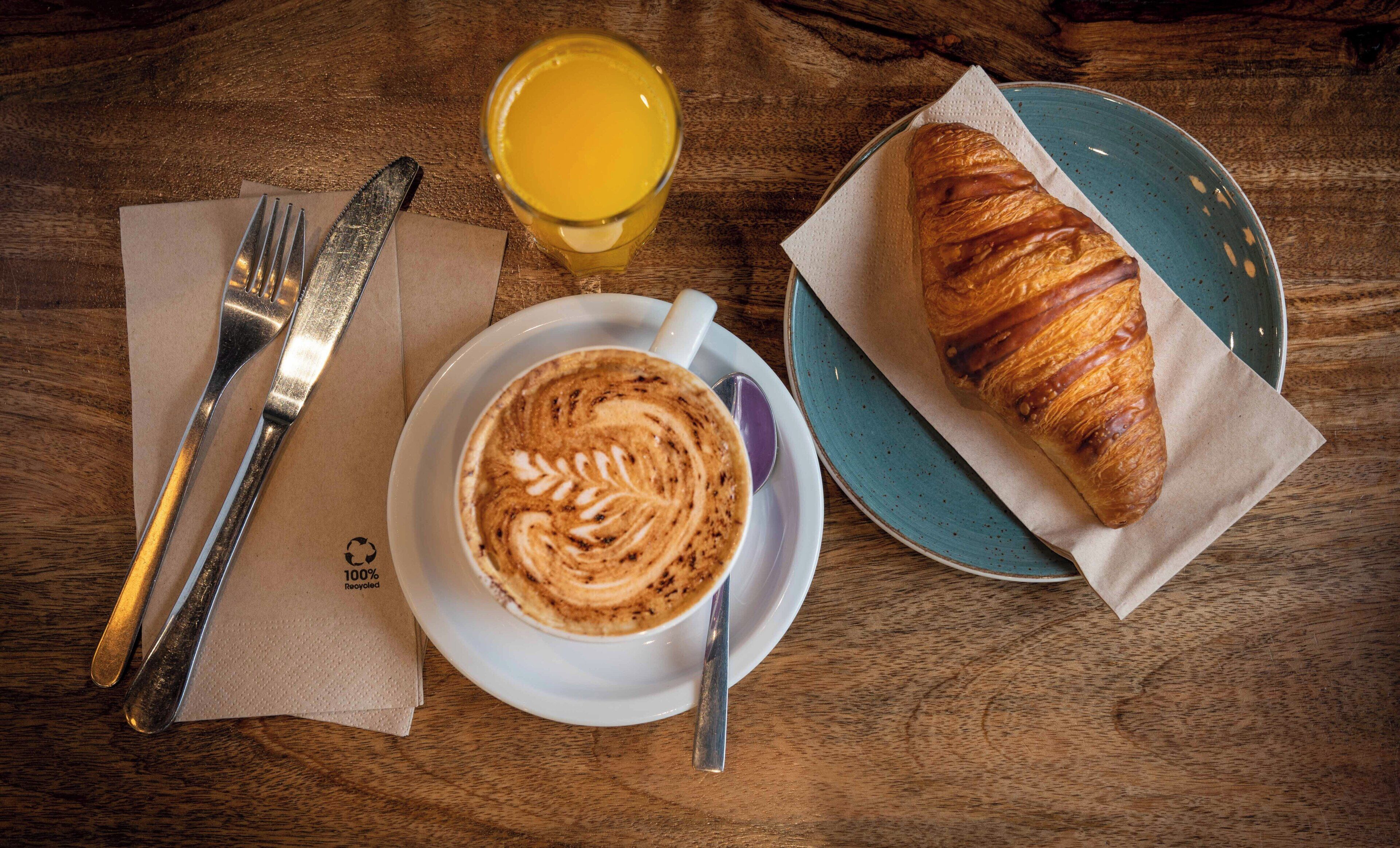 En frukostuppsättning med en croissant, en kopp kaffe och ett glas apelsinjuice på ett träbord.