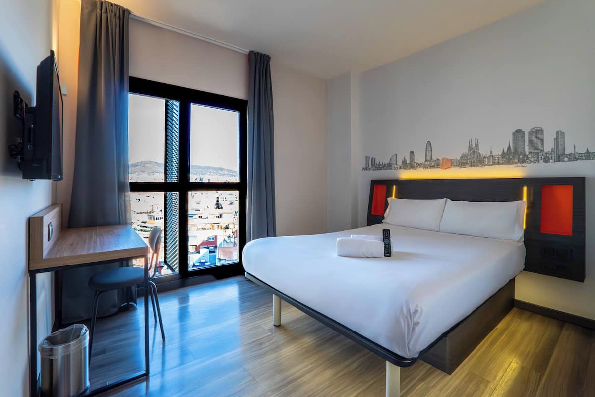 Habitación de hotel con cama, escritorio, silla, televisor, cuadro de paisaje urbano y vista a través de un ventanal grande.