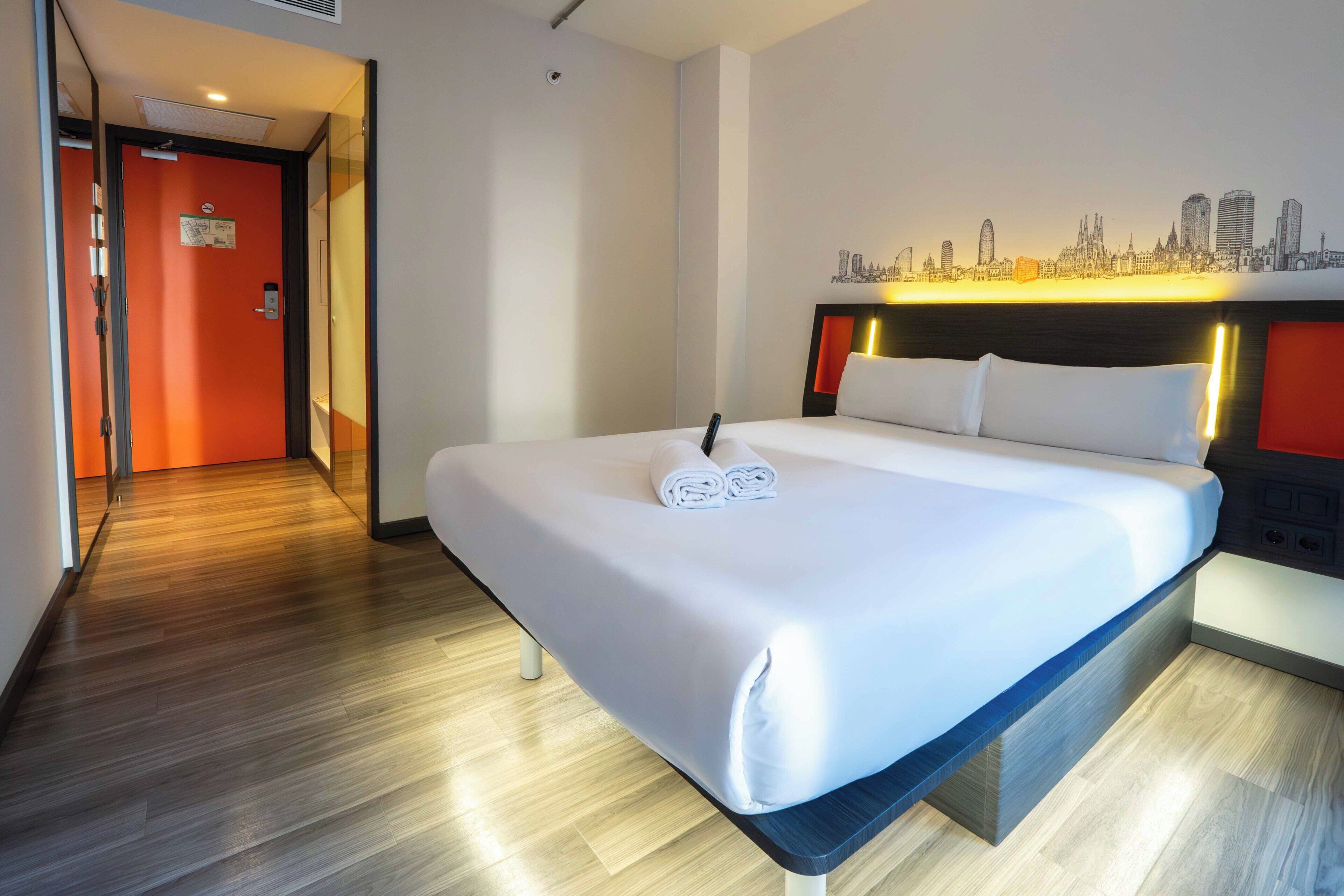A hotel room with a bed, a cityscape wall mural, a wooden floor, and a door with an orange wall.