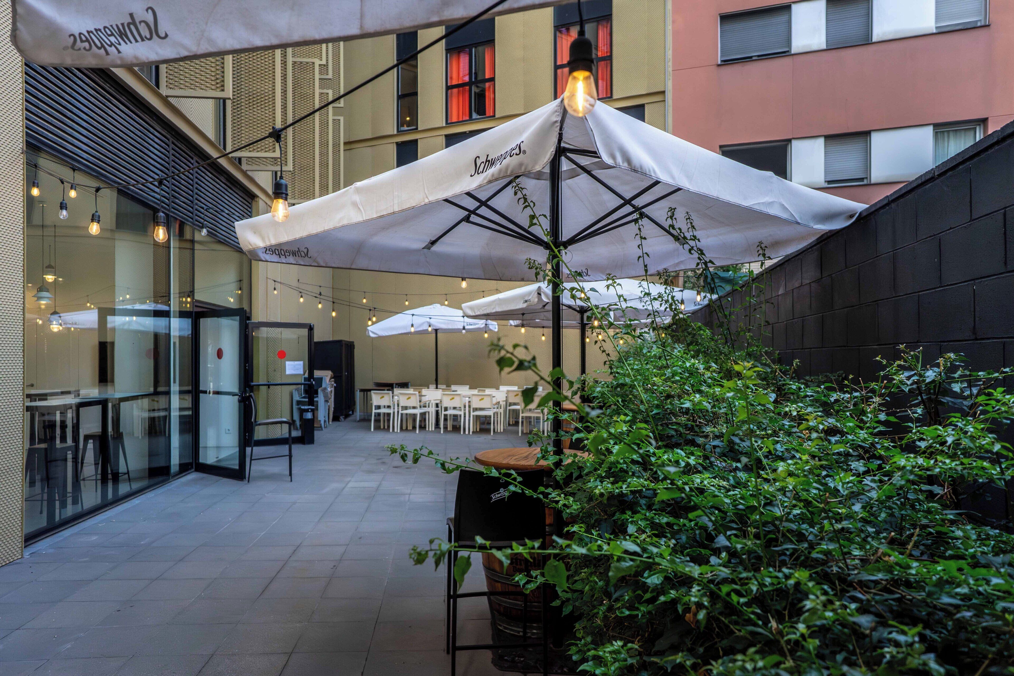 Een terras met parasols, tafels en stoelen, omringd door groen en een gevel.