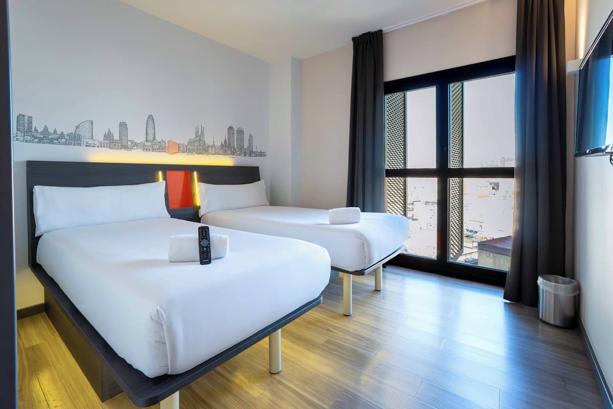 Habitación de hotel con dos camas, un mural de paisaje urbano en la pared, un ventanal con vistas y un cubo de basura.