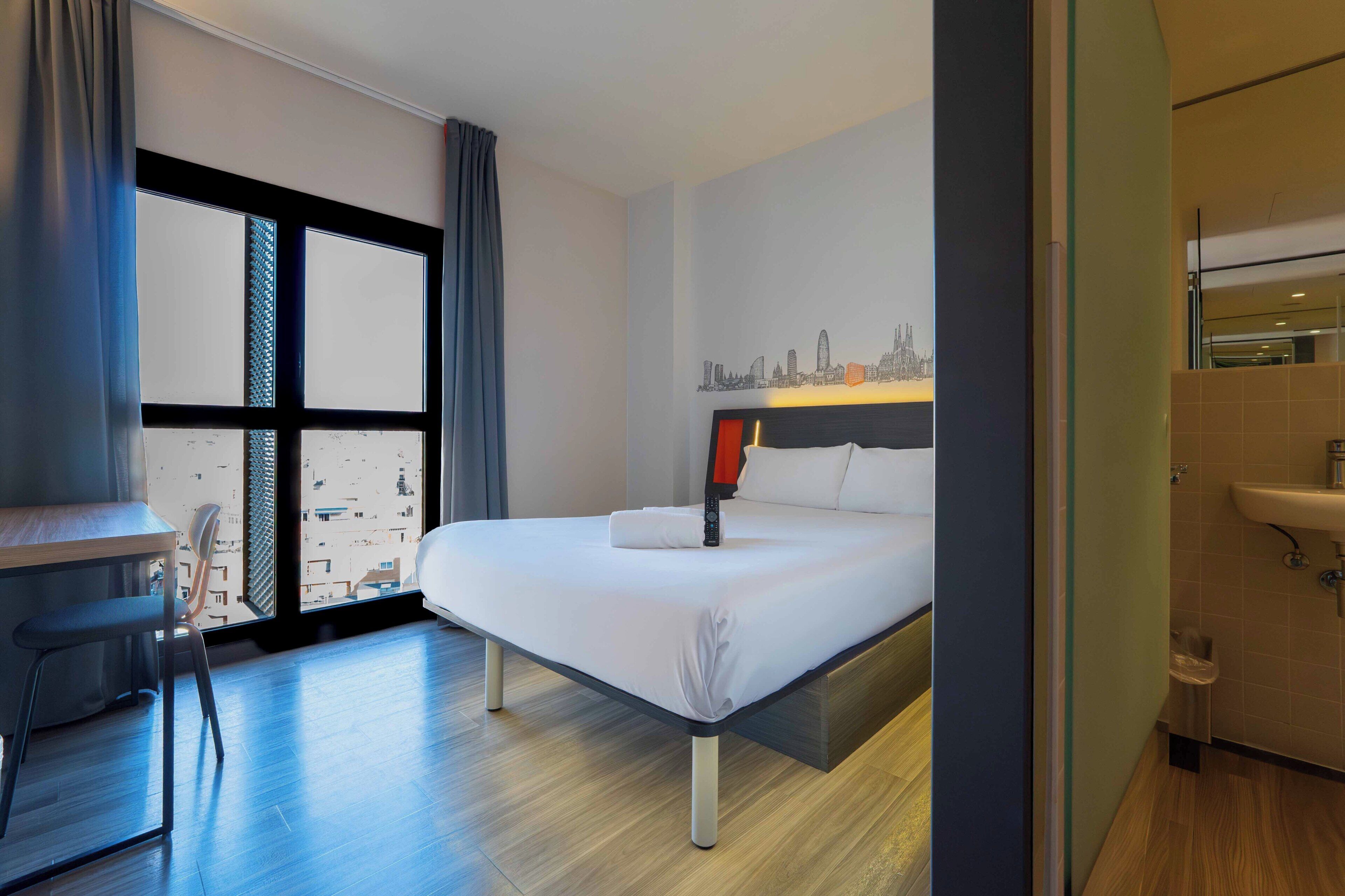 A hotel room with a bed, a desk, a chair, a window with curtains, and a cityscape view.