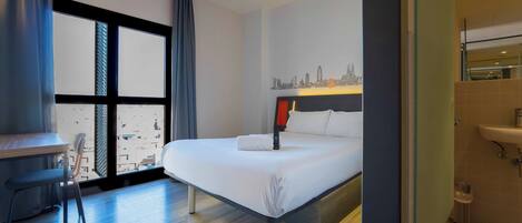 A hotel room with a bed, a desk, a chair, a window with curtains, and a cityscape view.