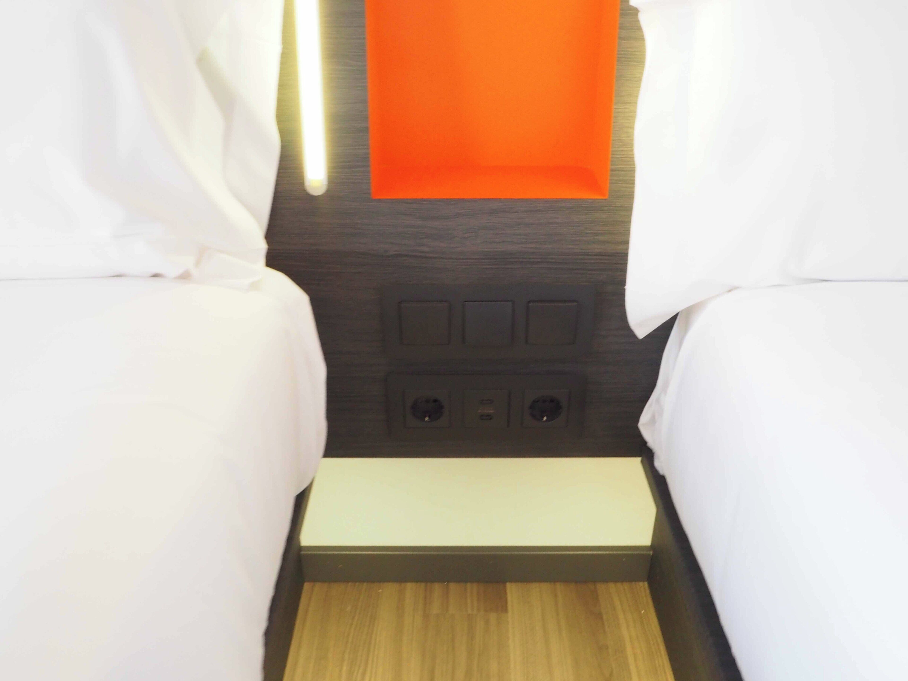 A hotel room with two beds, a nightstand, and a wall-mounted orange light fixture.