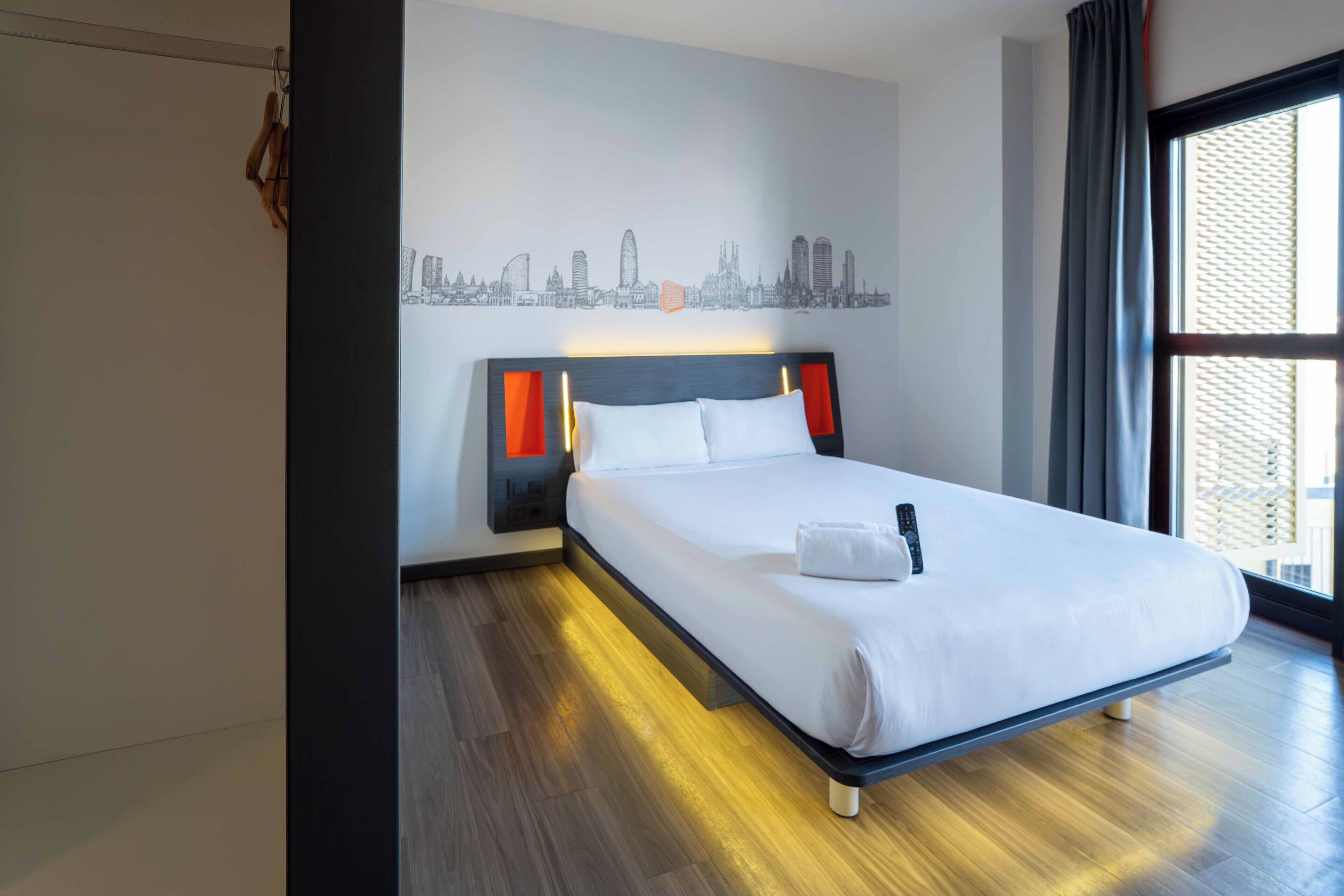 A hotel room with a bed, a cityscape wall art, a window with curtains, and a wooden floor.