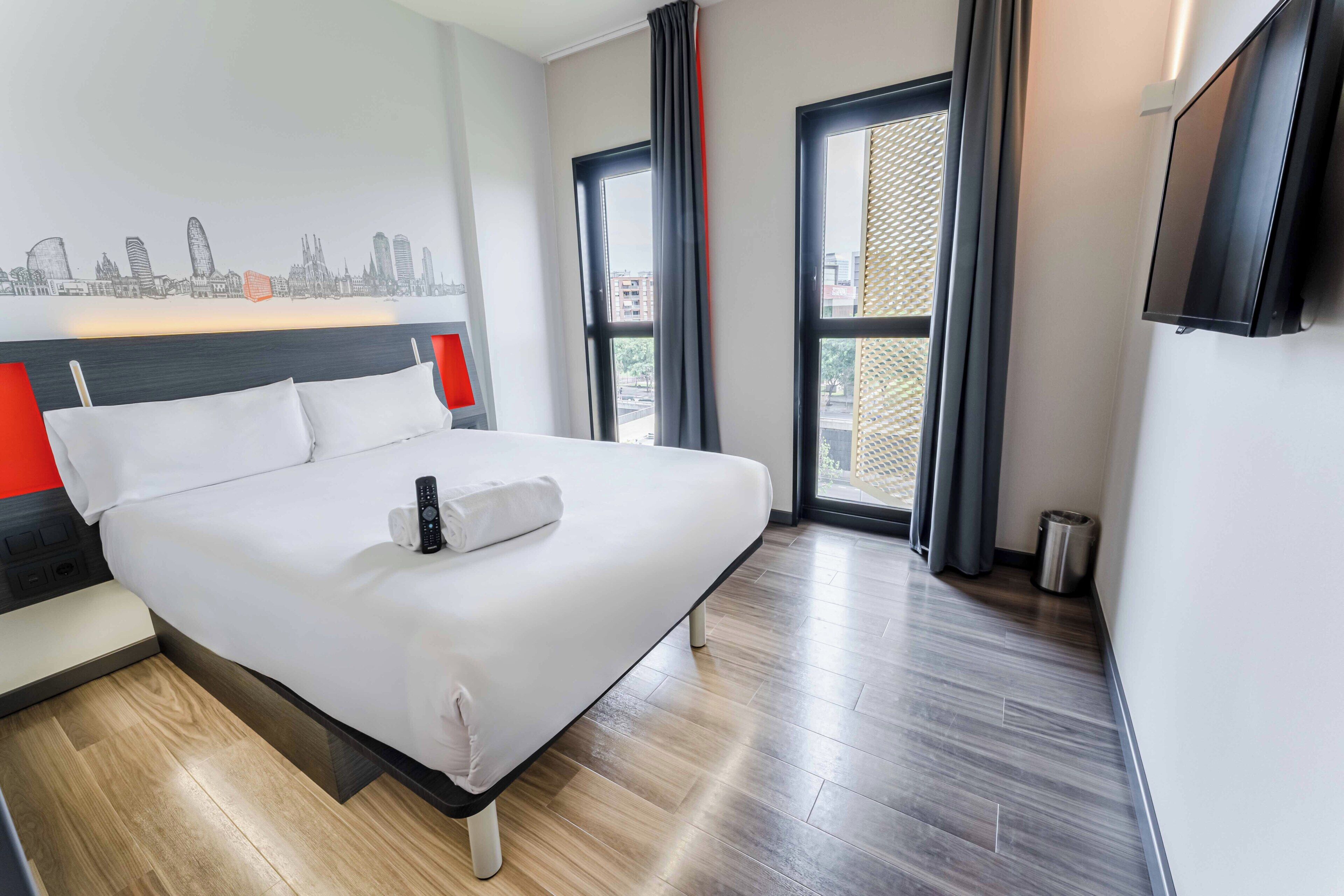 A hotel room with a bed, a cityscape mural, a TV, and a window with curtains.