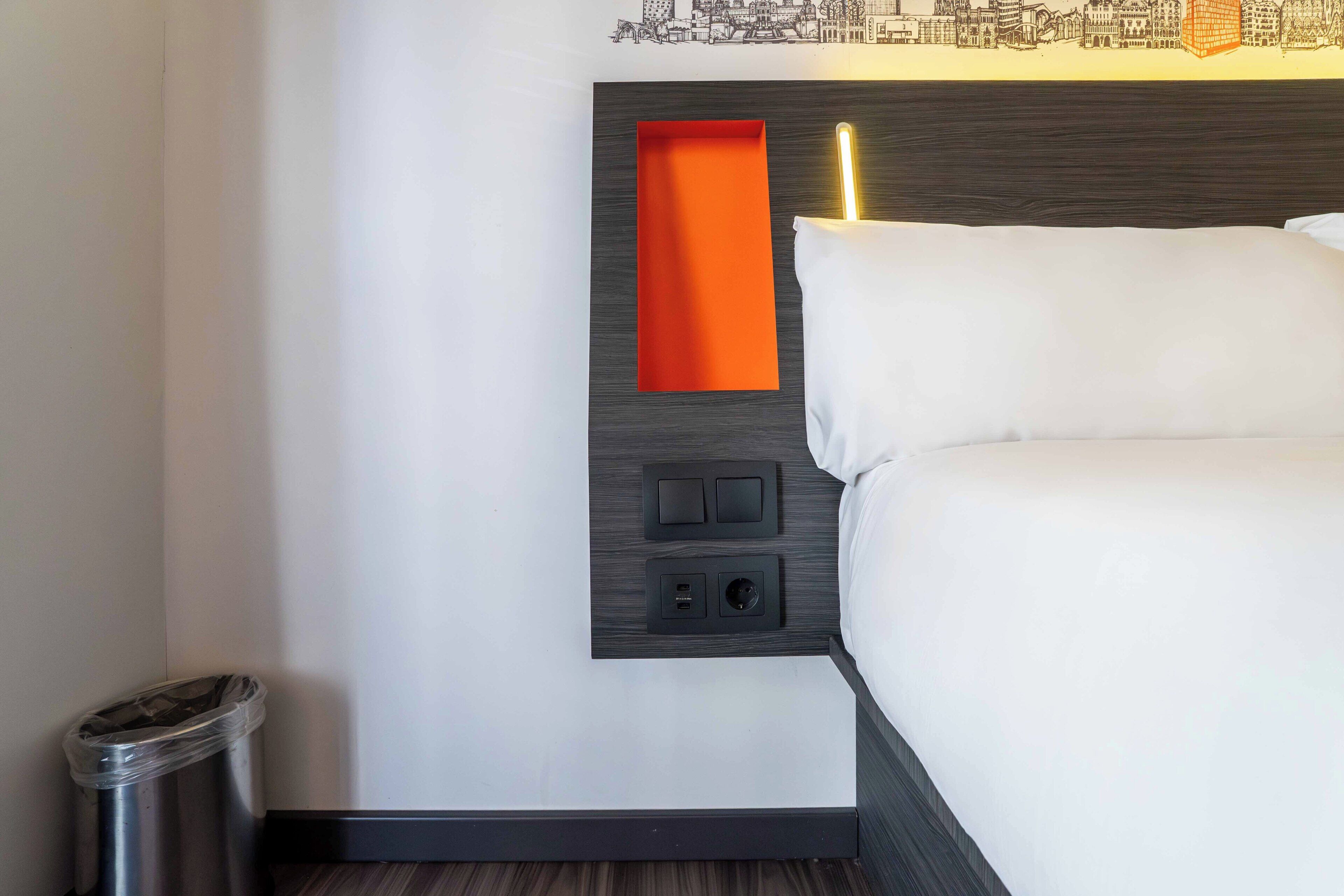 A hotel room with a bed, a trash bin, a wall-mounted power outlet, and a small orange shelf.