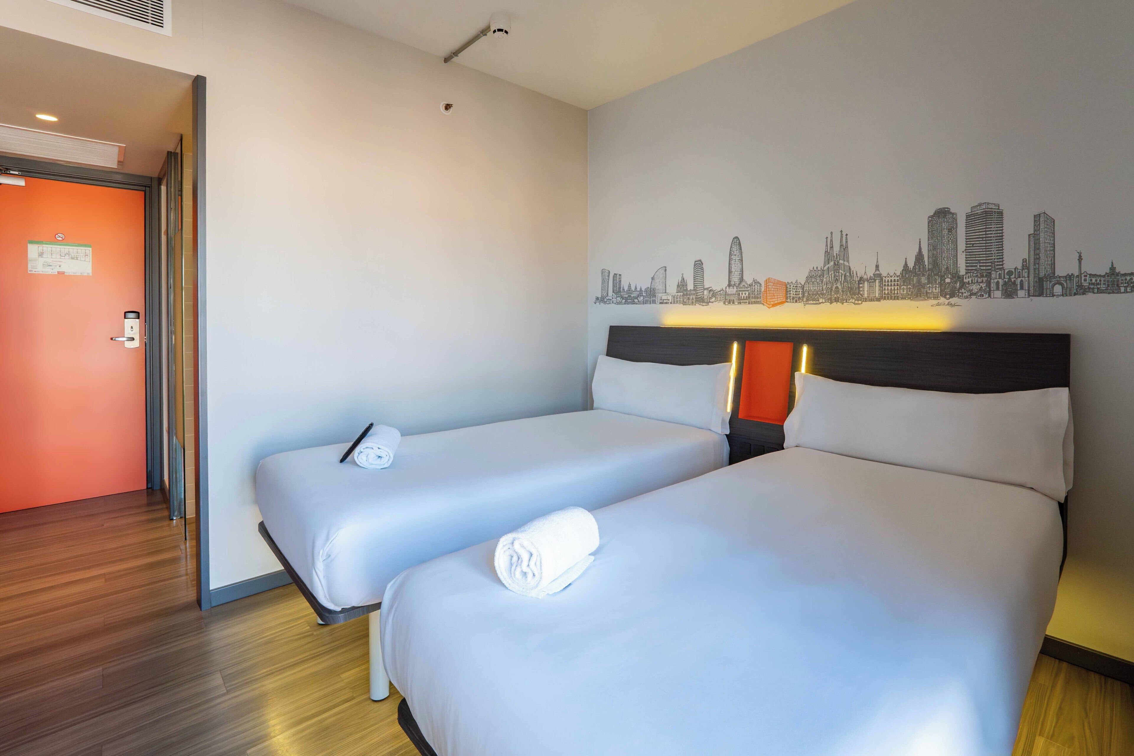 Hotellihuone, jossa on kaksi sänkyä, seinämaalaus, joka esittää kaupungin siluettia, ja ovi, jonka vieressä on oranssi seinä.