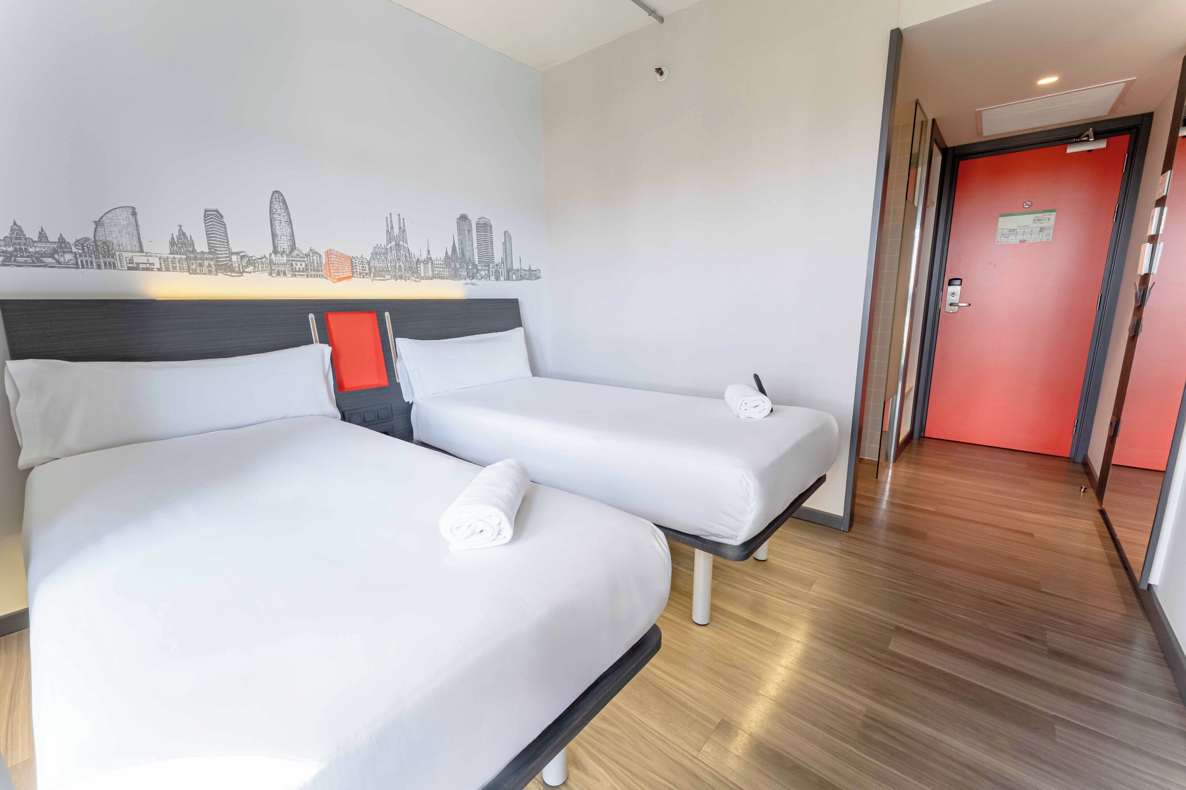A hotel room with two beds, a cityscape mural, a red door, and a wooden floor.