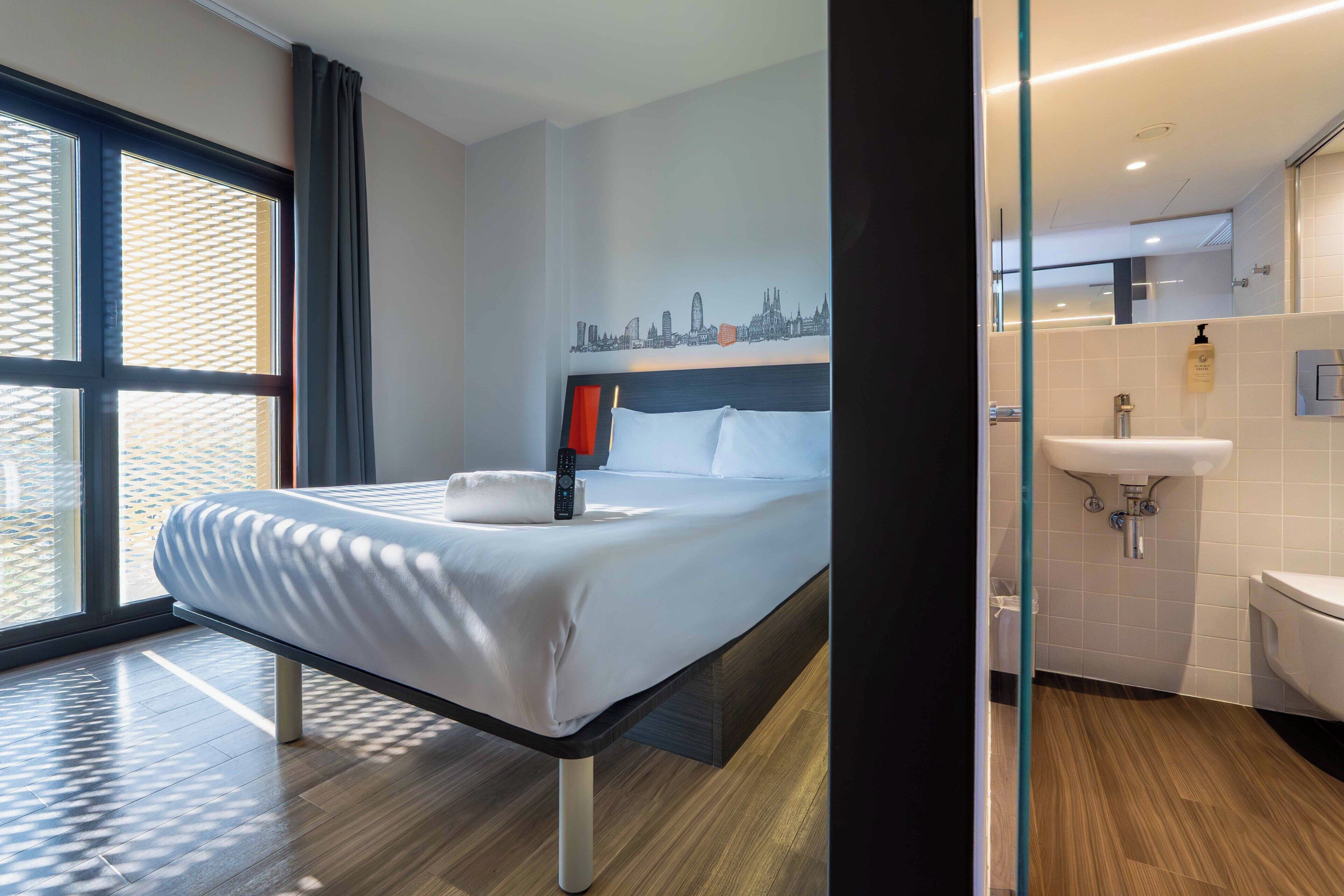 Habitación de hotel con una cama, un mural de la ciudad, un baño con lavamanos e inodoro, y ventanas amplias.