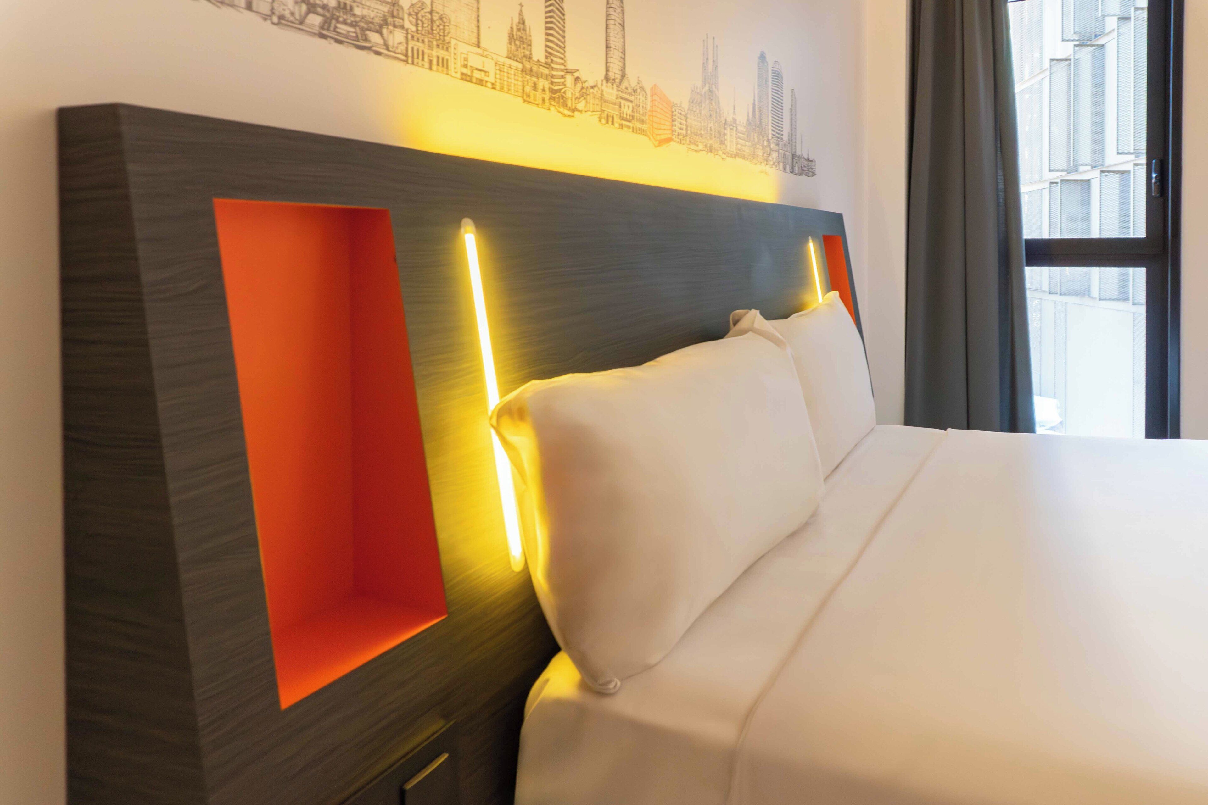 Habitación de hotel con cama, cabecera con iluminación integrada y un mural de paisaje urbano en la pared.