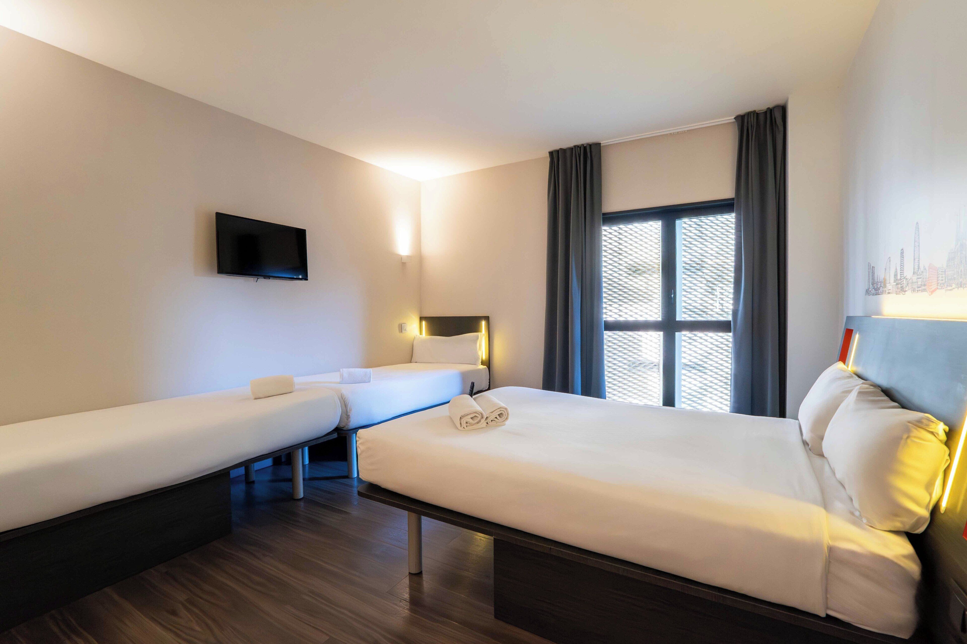 A hotel room with a large bed, a smaller bed, a TV, and a window with curtains.