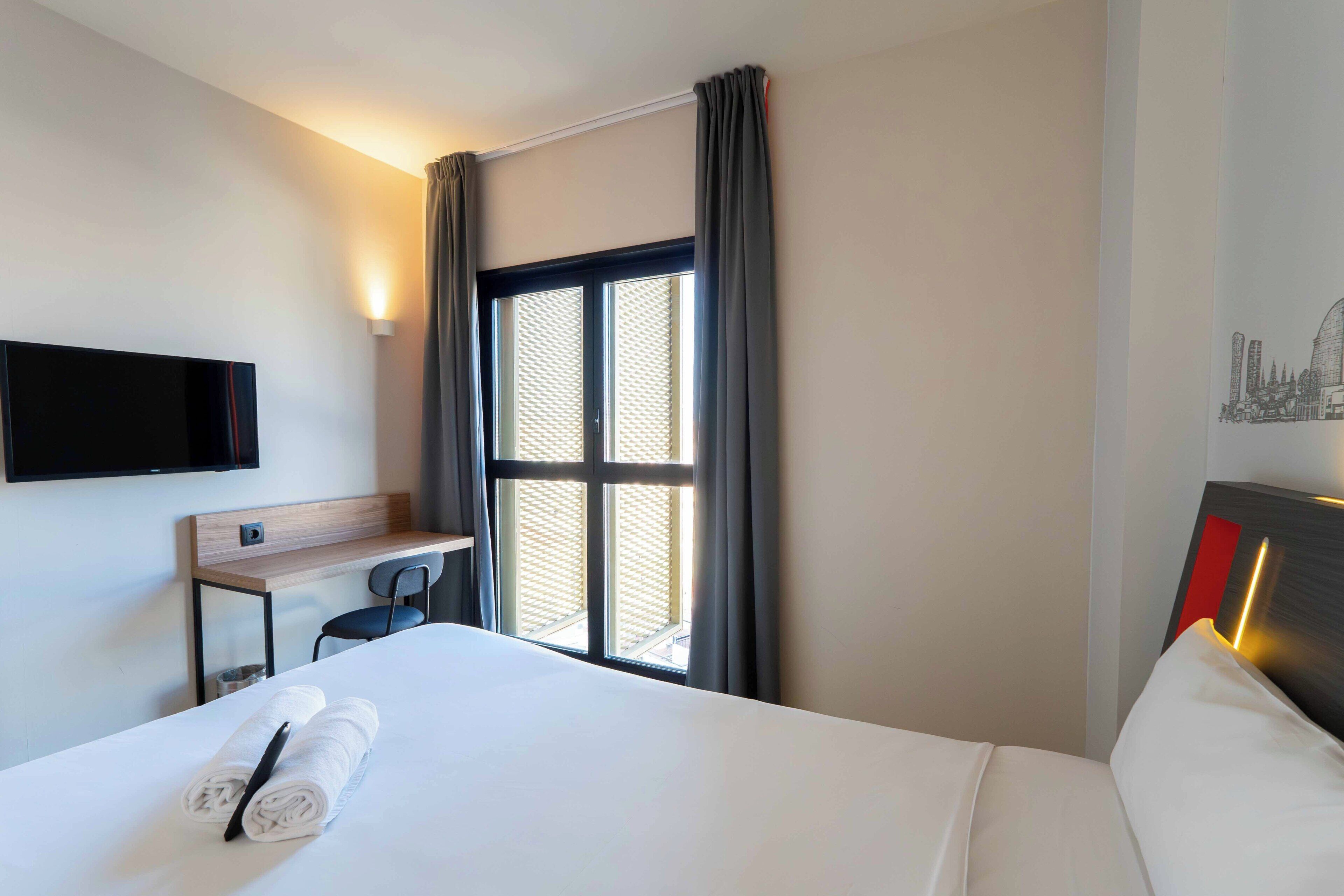 Una camera d'albergo con un letto, una scrivania, una sedia, una televisione e una finestra con tende.
