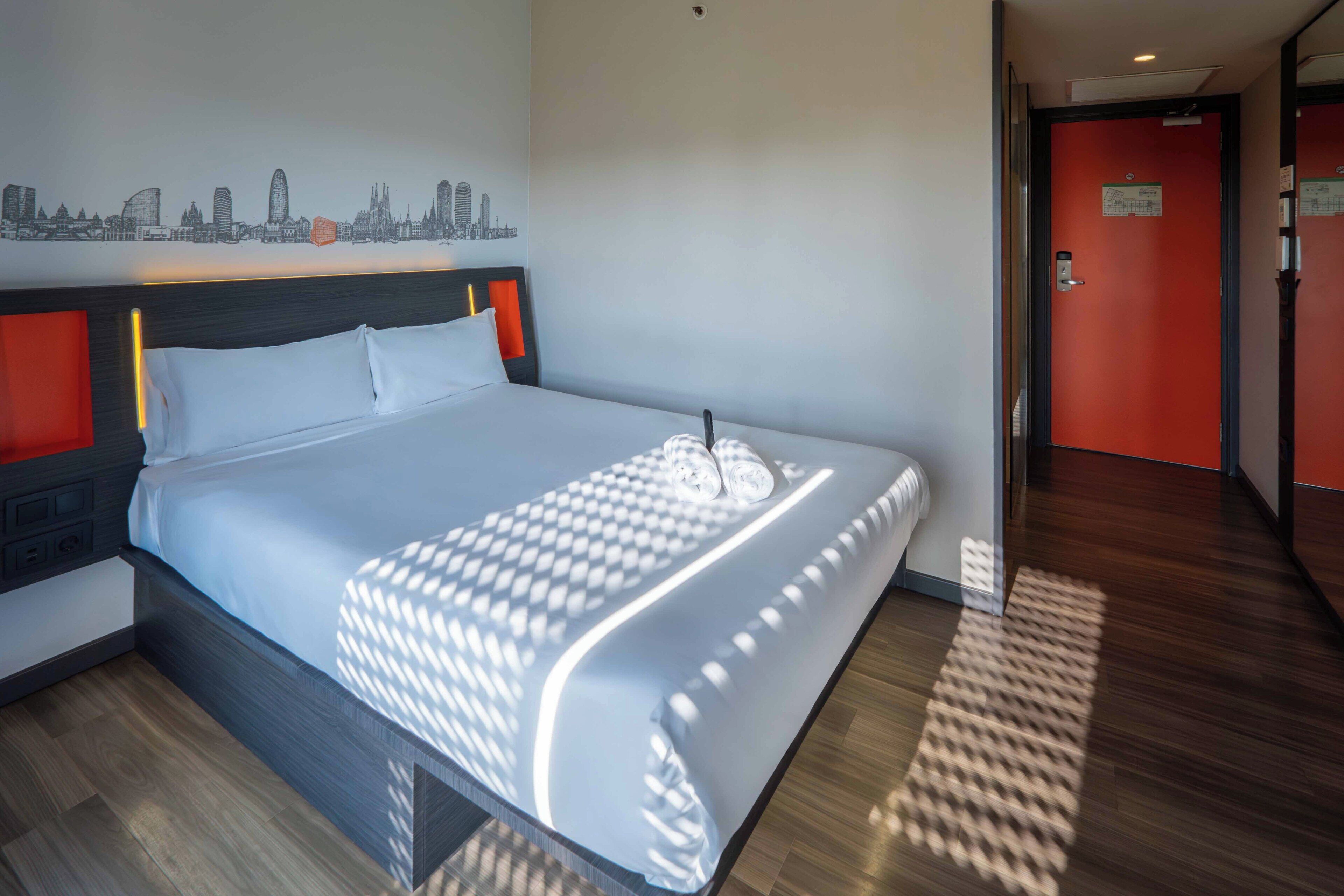 A hotel room with a bed, a cityscape wall art, a red door, and a patterned rug.