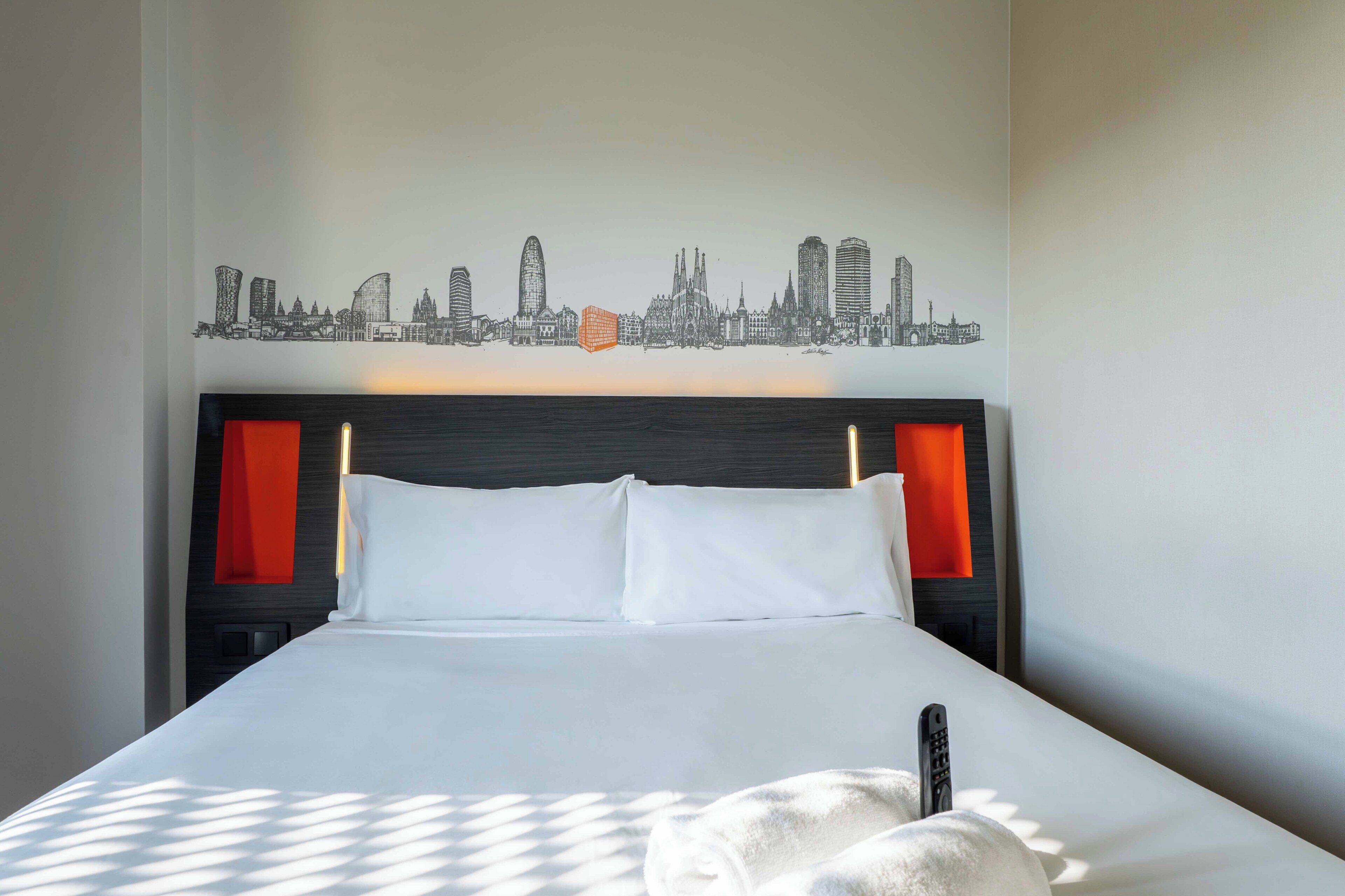 Une chambre d’hôtel avec un lit, une fresque représentant le panorama urbain sur un mur, et un mur d’accent de couleur rouge.