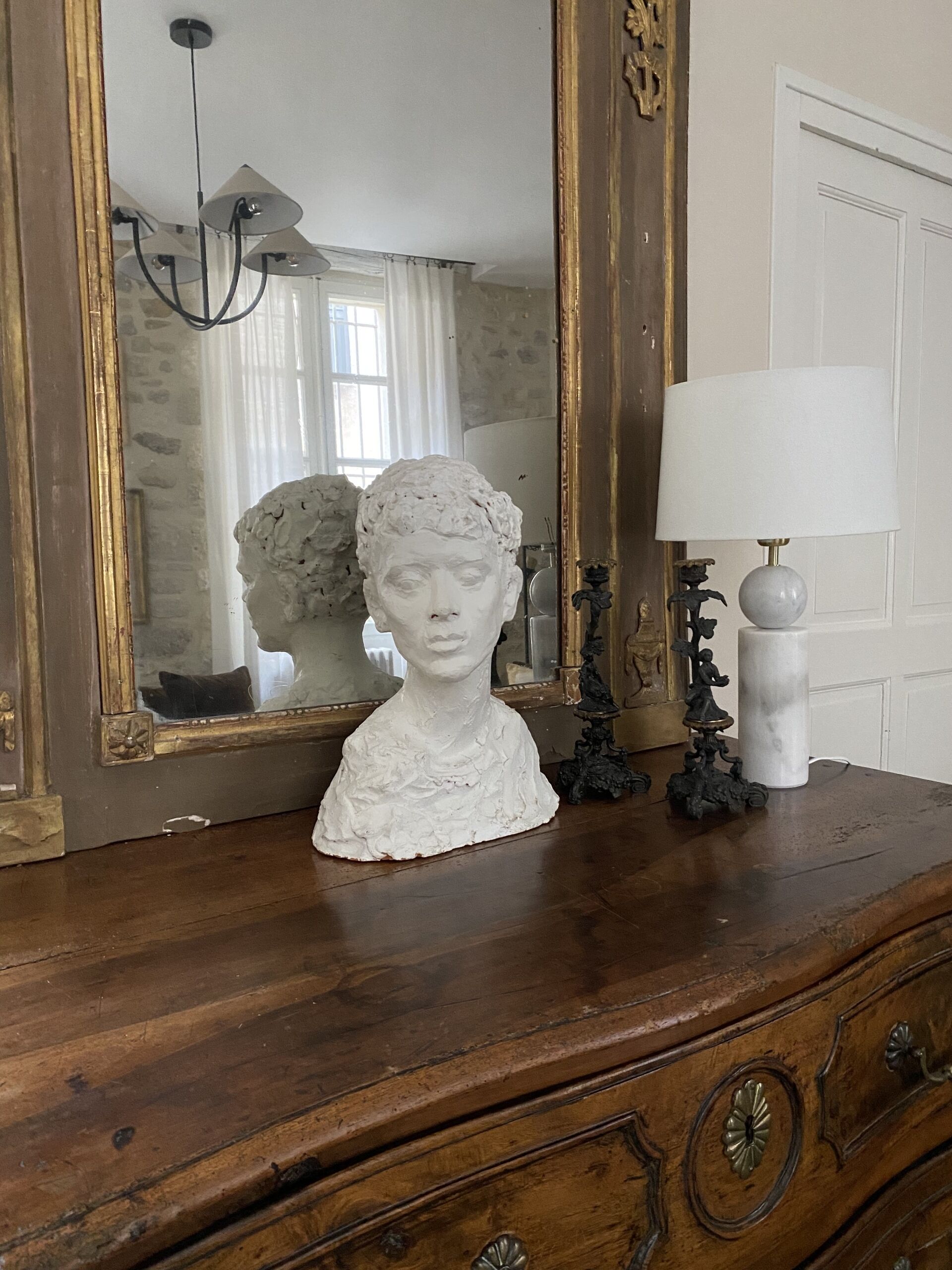 A wooden dresser with a white bust sculpture, a mirror, and a lamp.