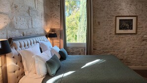 Een slaapkamer met een stenen muur, een groot raam met uitzicht op bomen, een bed met wit en groen beddengoed, en een lijst met een afbeelding aan de muur.
