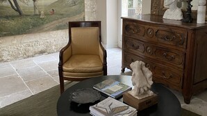 Een kamer met een groot landschapsprent, een houten dressoir, een stoel en een tafel met boeken en een beeldhouwwerk.