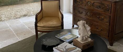 Una habitación con un cuadro de paisaje grande, un aparador de madera, una silla y una mesa con libros y una escultura.