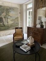Una habitación con un cuadro de paisaje grande, un aparador de madera, una silla y una mesa con libros y una escultura.