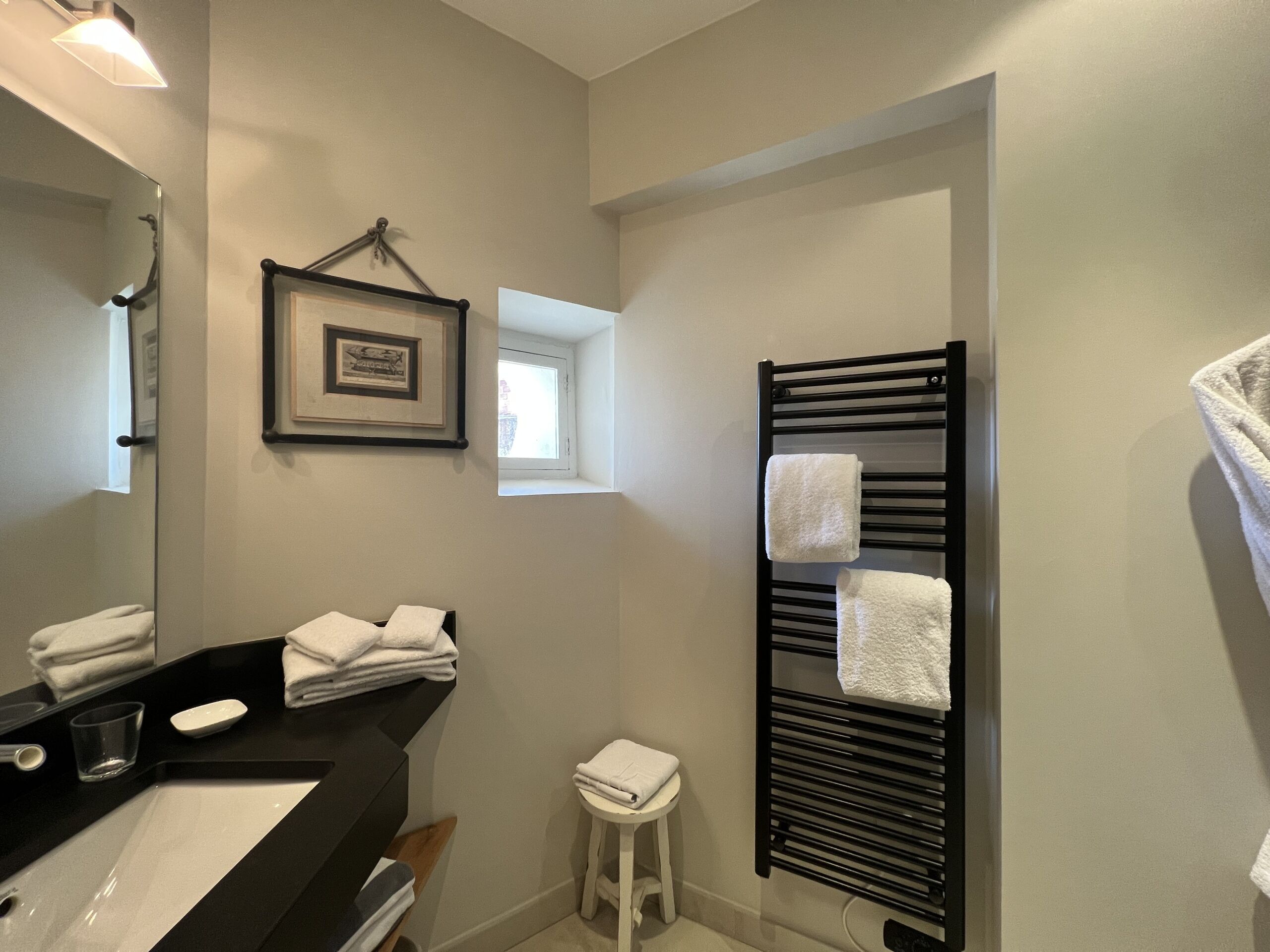 A bathroom with a black sink, white countertop, and a black towel rack with towels. There is a small window and a framed picture on the wall.