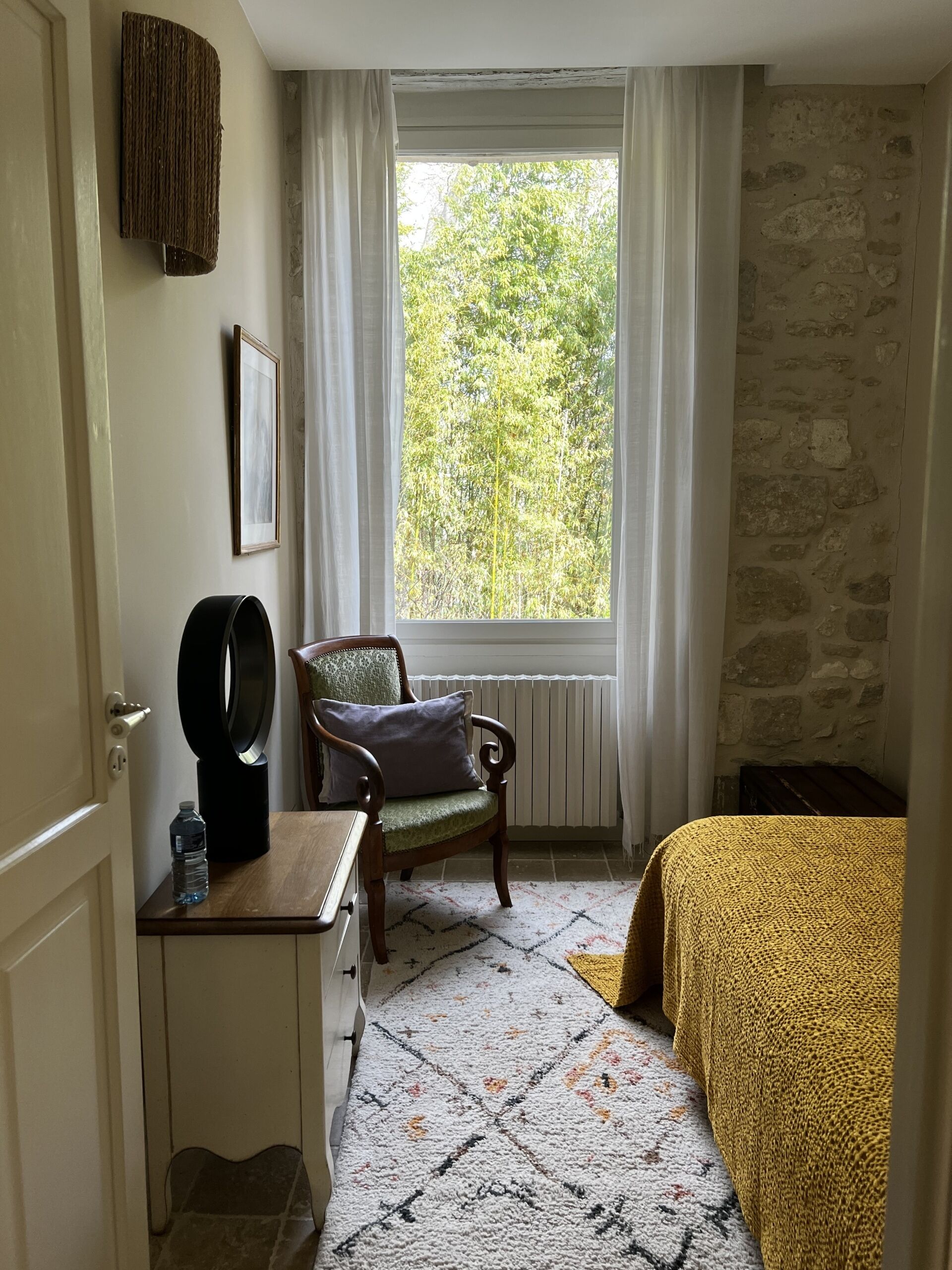A room with a bed, a chair, a radiator, a window with curtains, a door, and a wall with a stone pattern.