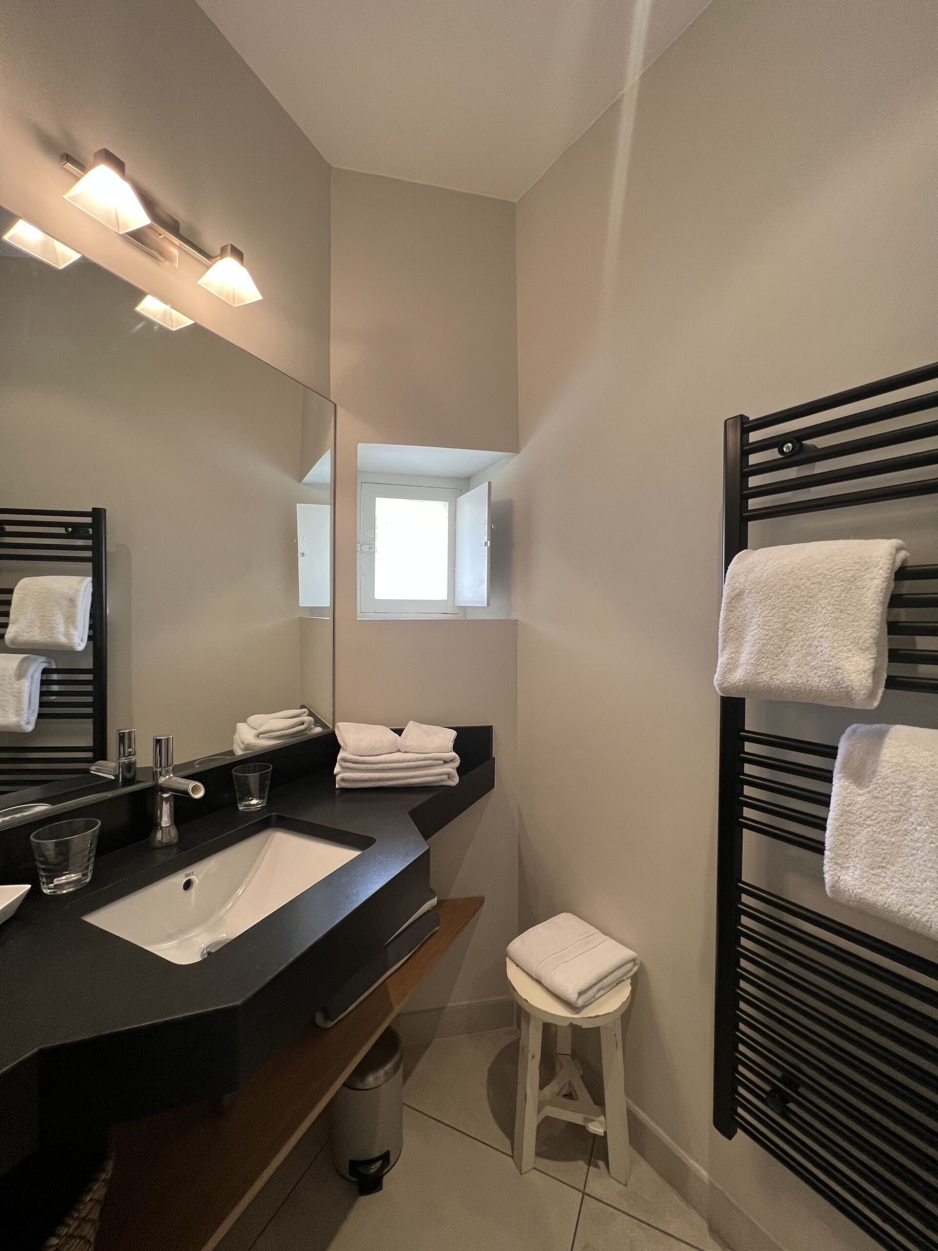 A modern bathroom with a black vanity, a white sink, and a mirror. There are towels on the wall and a small stool.