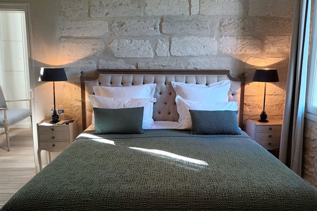 Een net opgemaakt bed met een groene deken, twee witte kussens en twee groene kussens. Twee nachtkastjes met lampen, een stoel en een stenen muur achter het bed.