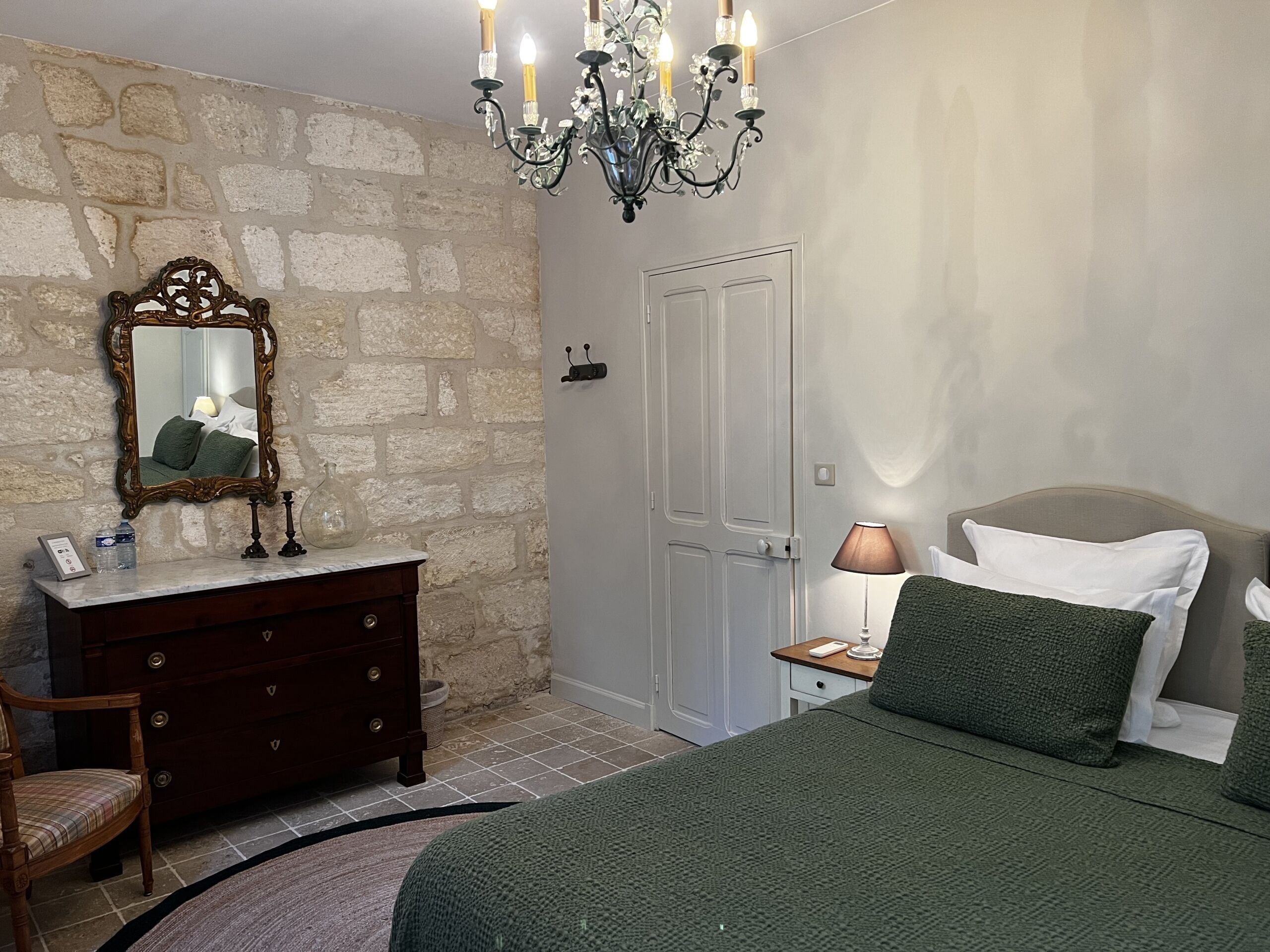 A bedroom with a stone wall, a wooden dresser, a bed with green bedding, a chandelier, and a mirror.