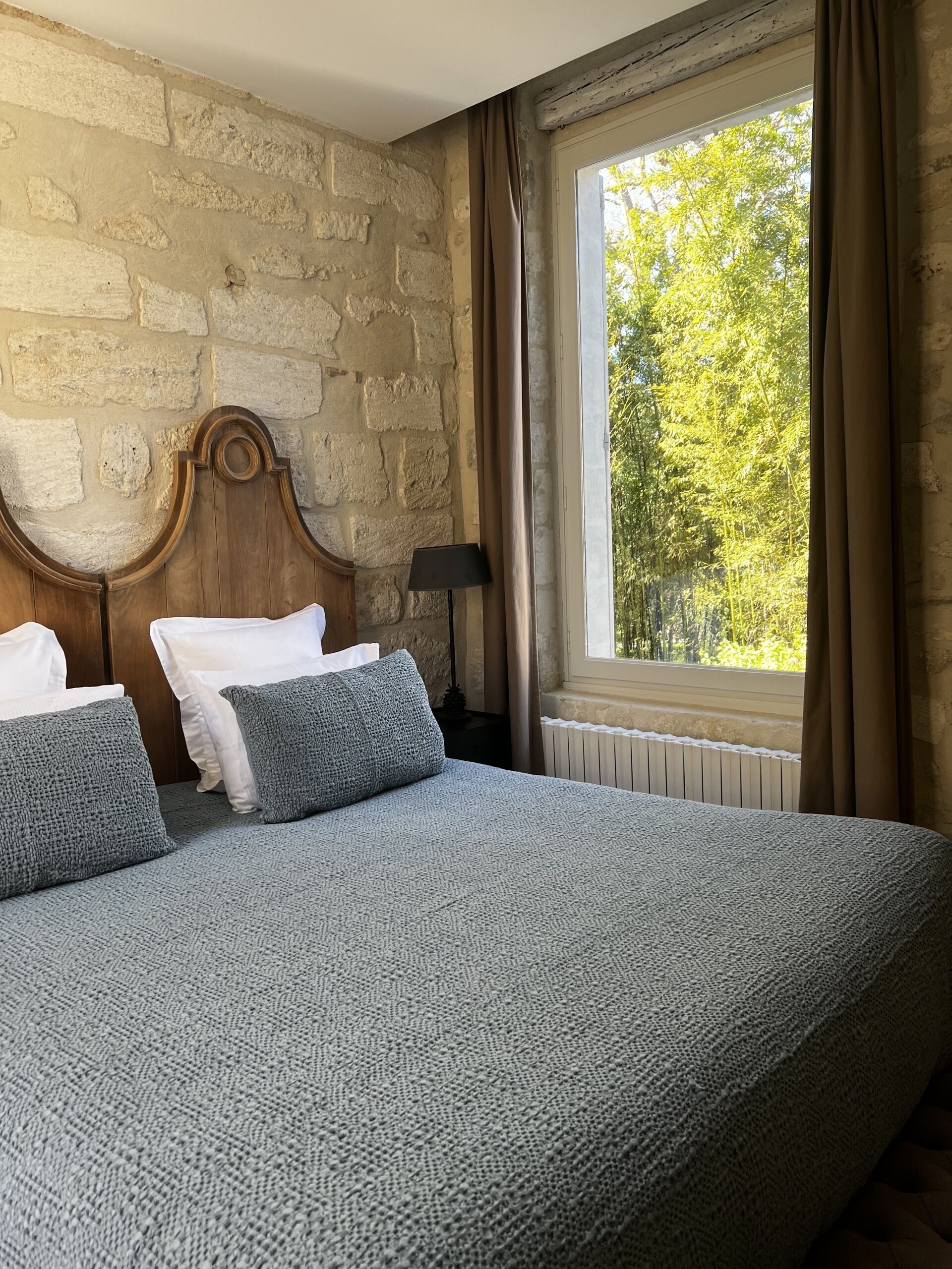 A bedroom with a stone wall, a wooden headboard, a large window with curtains, and a bed with grey bedding and pillows.