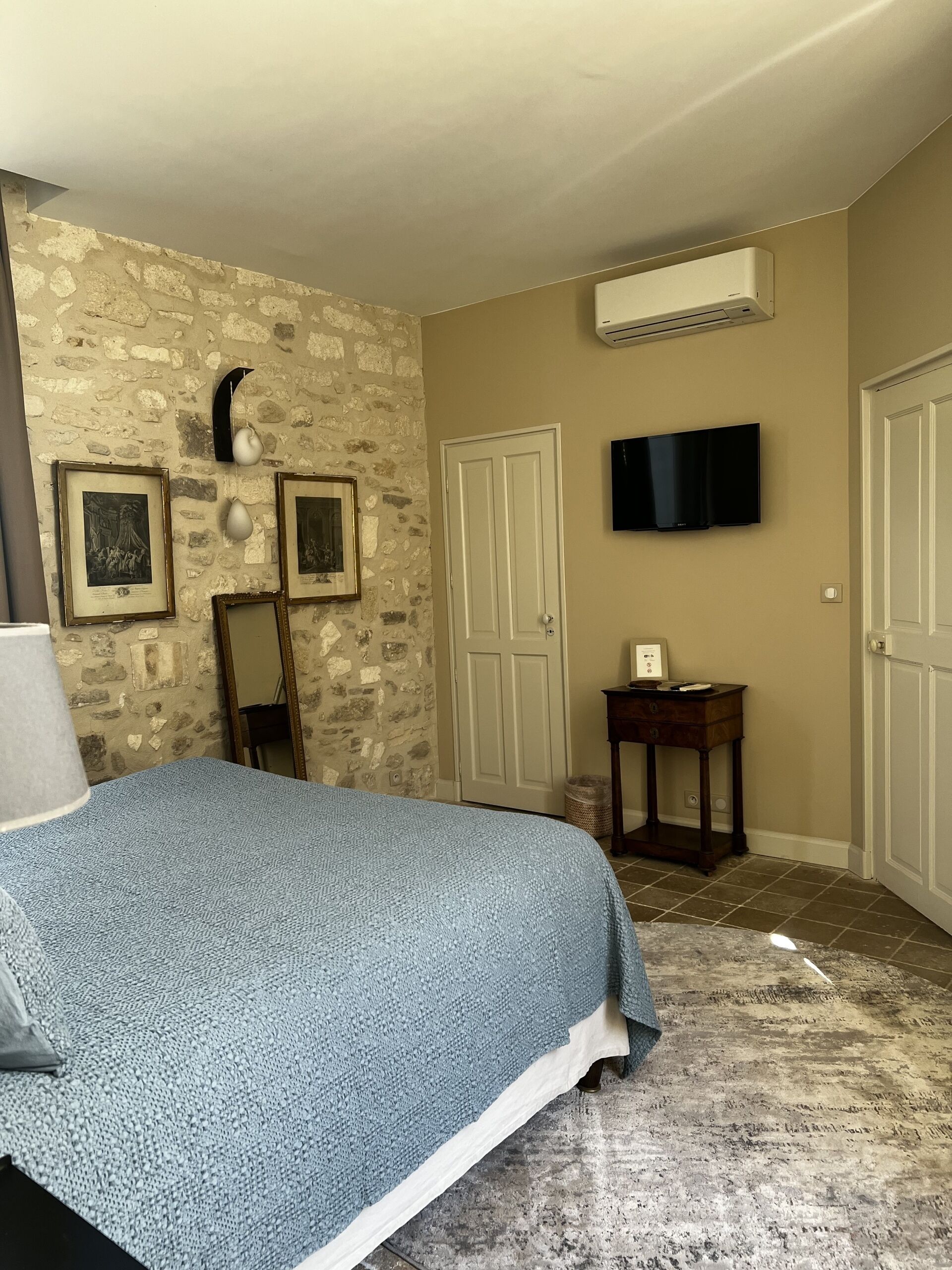 A bedroom with a stone wall, a bed with a blue blanket, a flat-screen TV, and a small table with a lamp.