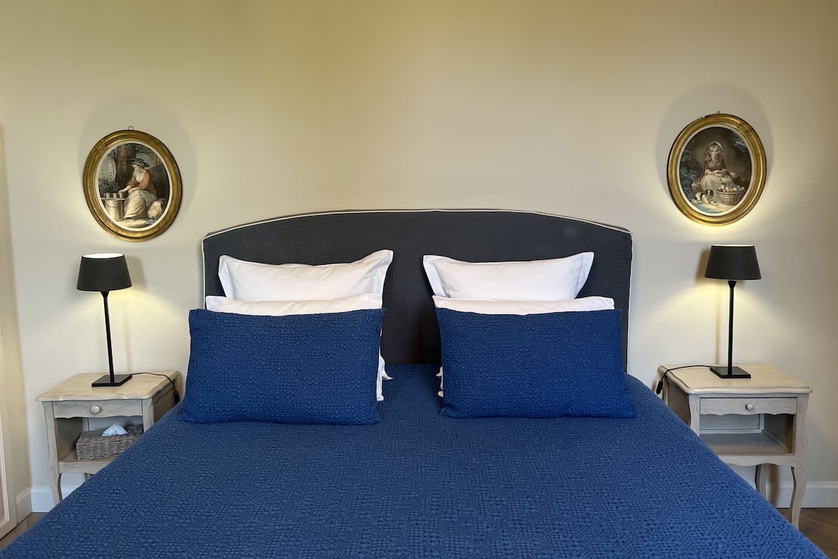 Een bed met een donker hoofdbord, twee witte kussens en twee blauwe kussens. Twee nachtkastjes met lampen en ingekaderde foto’s aan de muur.
