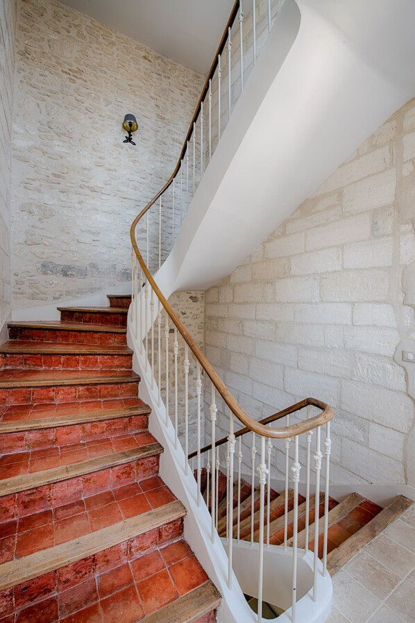 A staircase with red-tiled steps and white balusters, leading to a landing with a white wall and a decorative wall-mounted object. - N°15 Les Confidences - Chambres D'hôtes (Avignon)